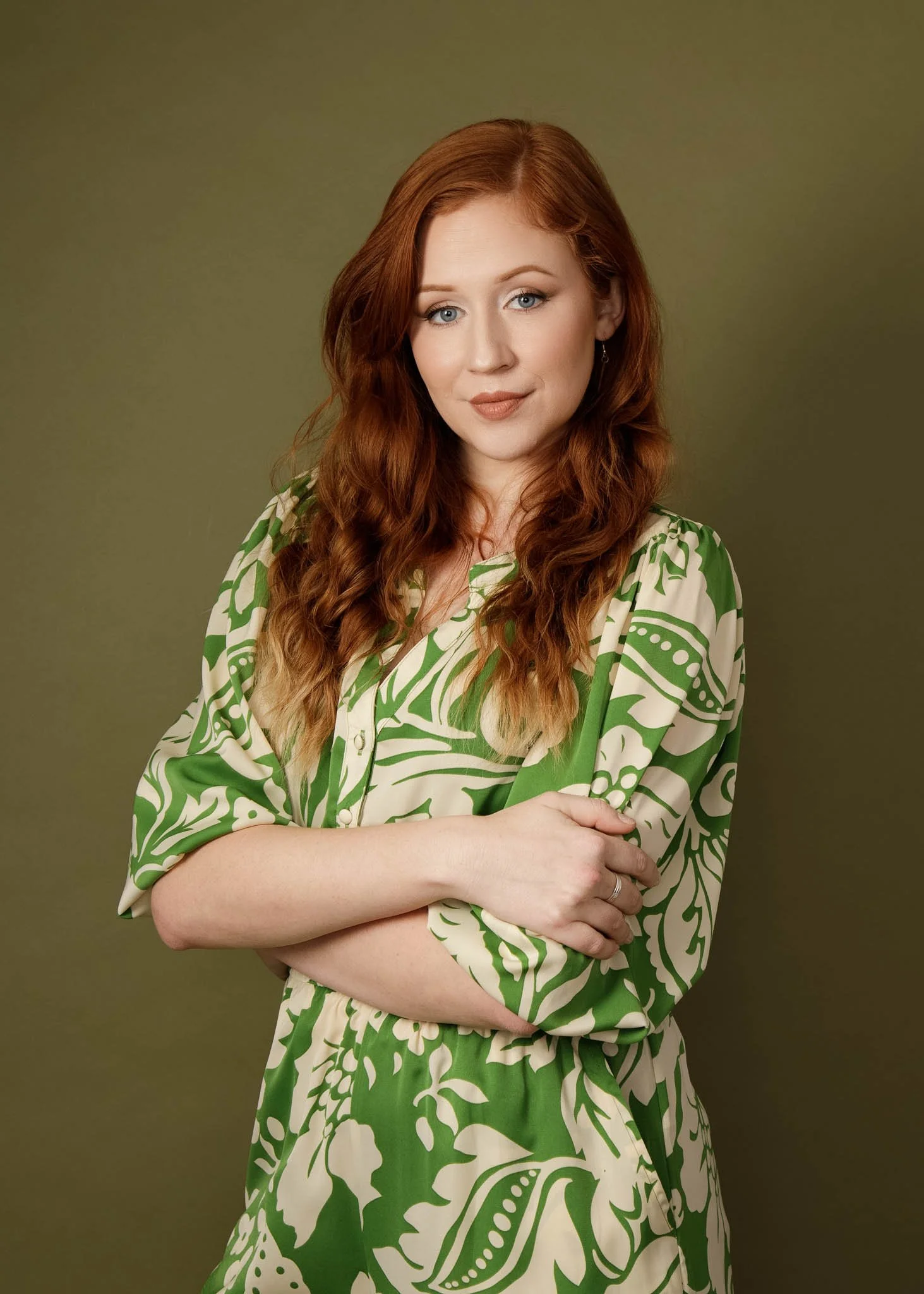 woman with long red hair and relaxed, natural expression, standing crossed arms posed, wearing green/cream print outfit on olive green backdrop in studio
