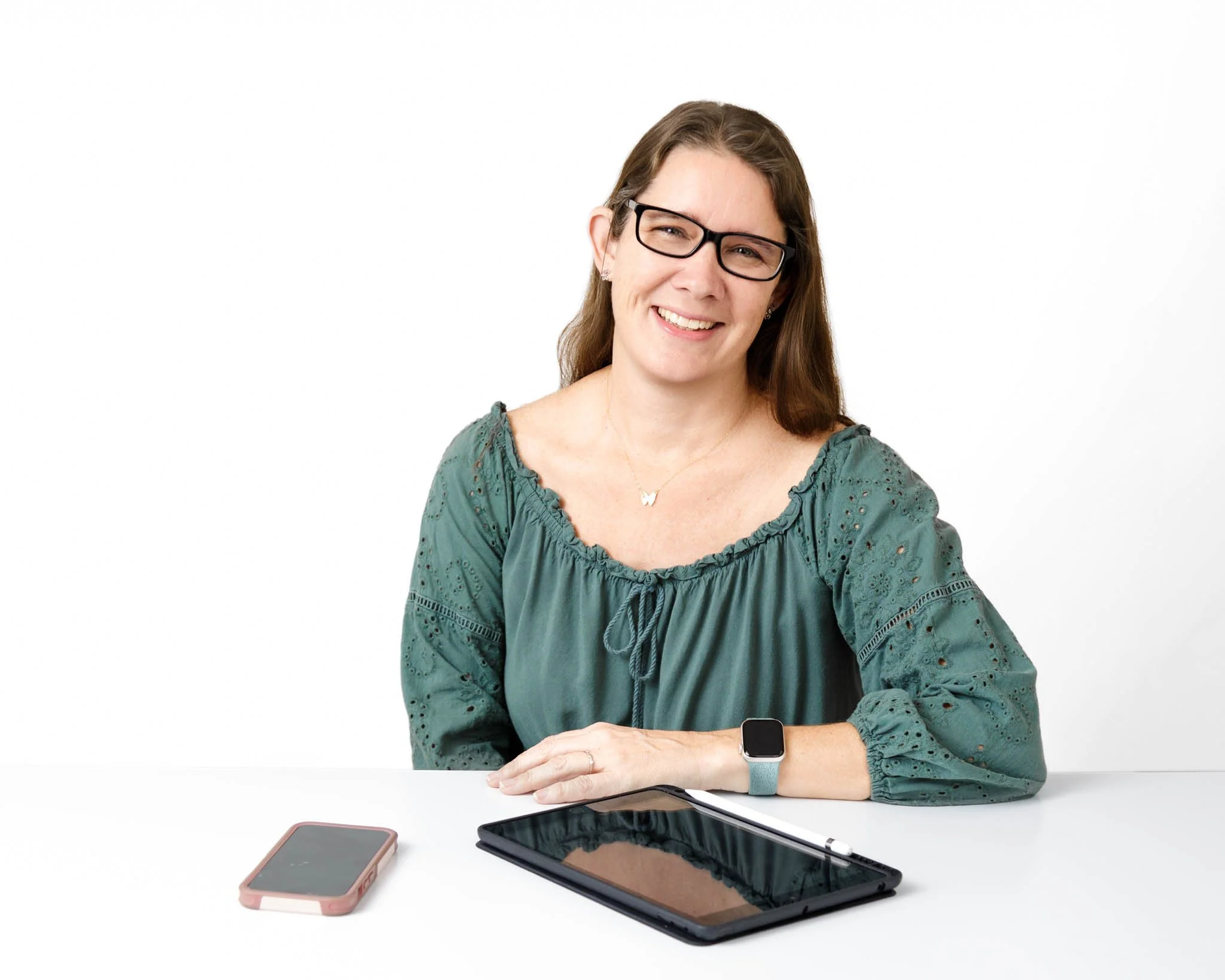 brand photo woman at desk with tablet and phone wearing glasses, green casual shirt and long brown hair on white backdrop / Photo by Erin Valkner Photography of New Braunfels, Texas. See more at www.erinvalknerphotography.com/blog