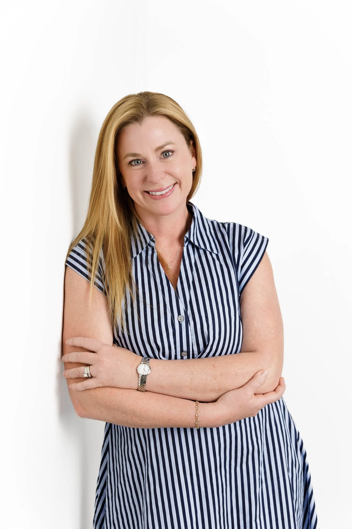 woman smiling with strawberry blond hair leaning against white wall with arms crossed wearing blue and white striped dress in studio / Photo by Erin Valkner Photography of New Braunfels, Texas. See more at www.erinvalknerphotography.com/blog