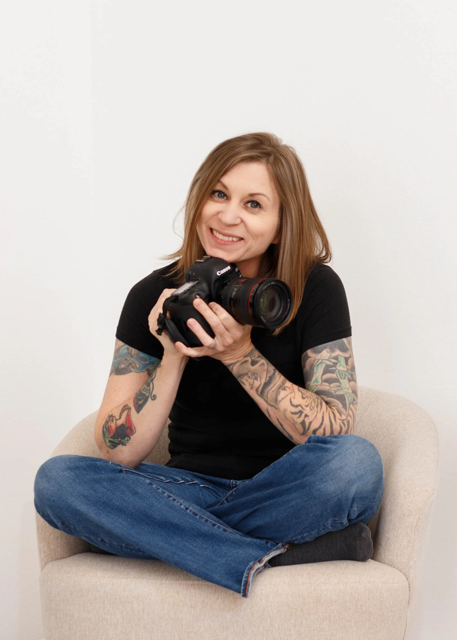 branding photo of female photographer holding camera seated cross legged in chair white backdrop / Photo by Erin Valkner Photography of New Braunfels, Texas. See more at www.erinvalknerphotography.com/blog