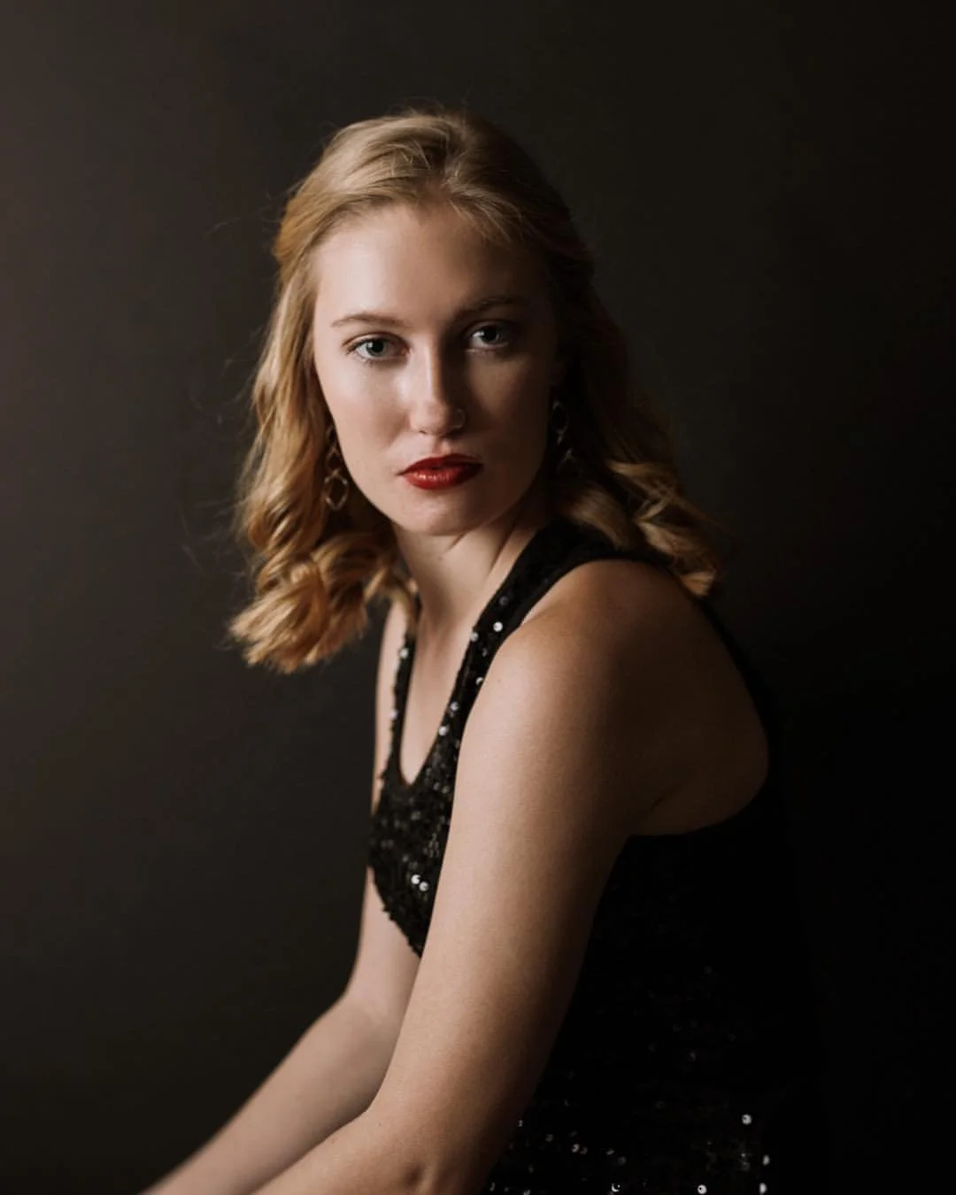 Studio portrait of woman with blond hair, intense look at camera with solemn expression, wearing black top with sequins and sparkles, pose seated in profile with face to camera