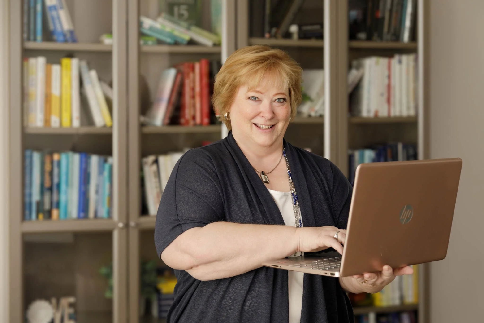 branding photo of woman standing in front of bookcase holding laptop looking at camera smiling / Photo by Erin Valkner Photography of New Braunfels, Texas. See more at www.erinvalknerphotography.com/blog