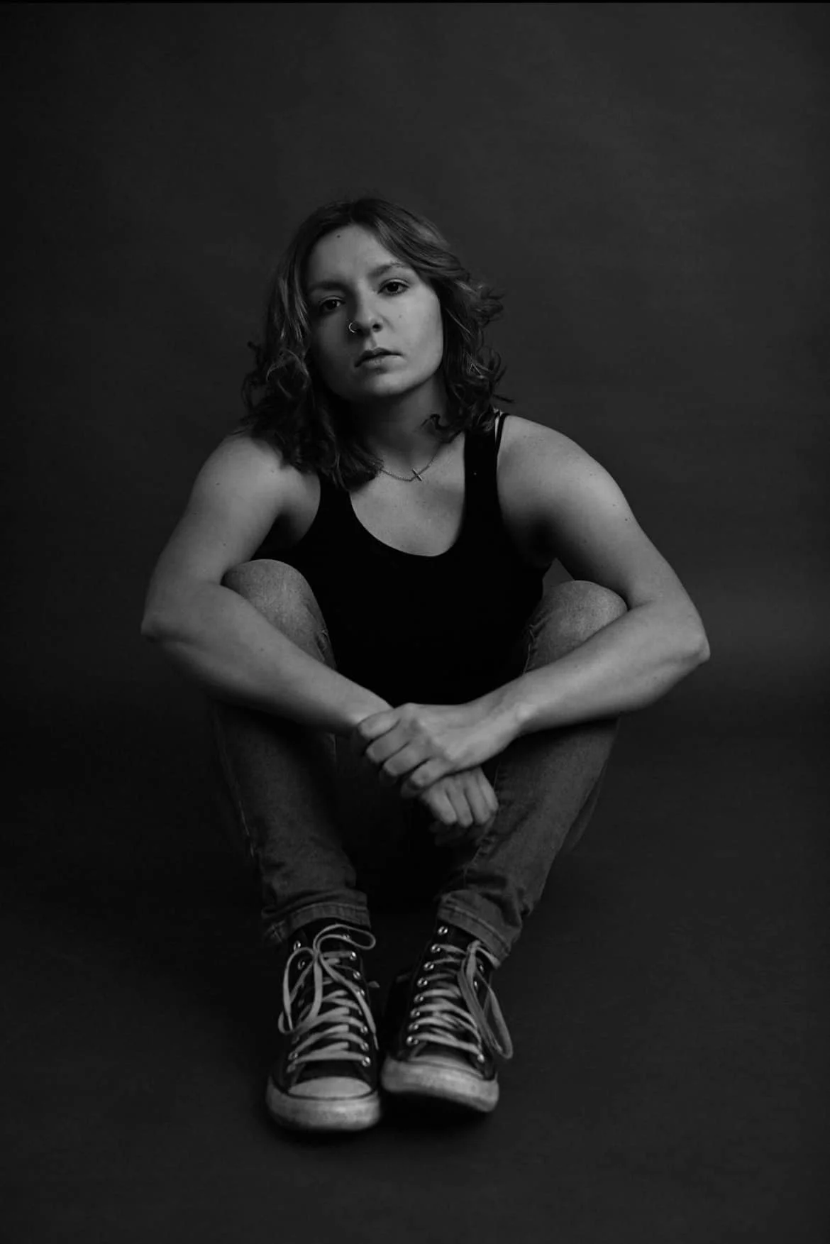 black and white image of woman with short hair, nose ring, necklace, wearing black tank top, jeans and converse, with neutral expression sitting in seated pose with arms around knees up