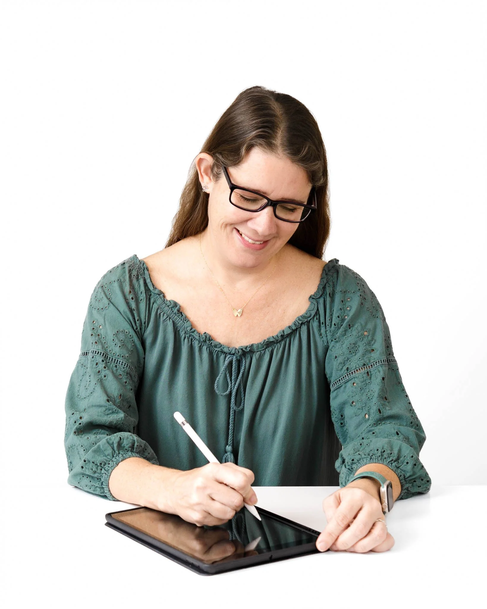 woman wearing glasses and green top long brown hair sitting at white desk writing with tablet and pen white background / Photo by Erin Valkner Photography of New Braunfels, Texas. See more at www.erinvalknerphotography.com/blog