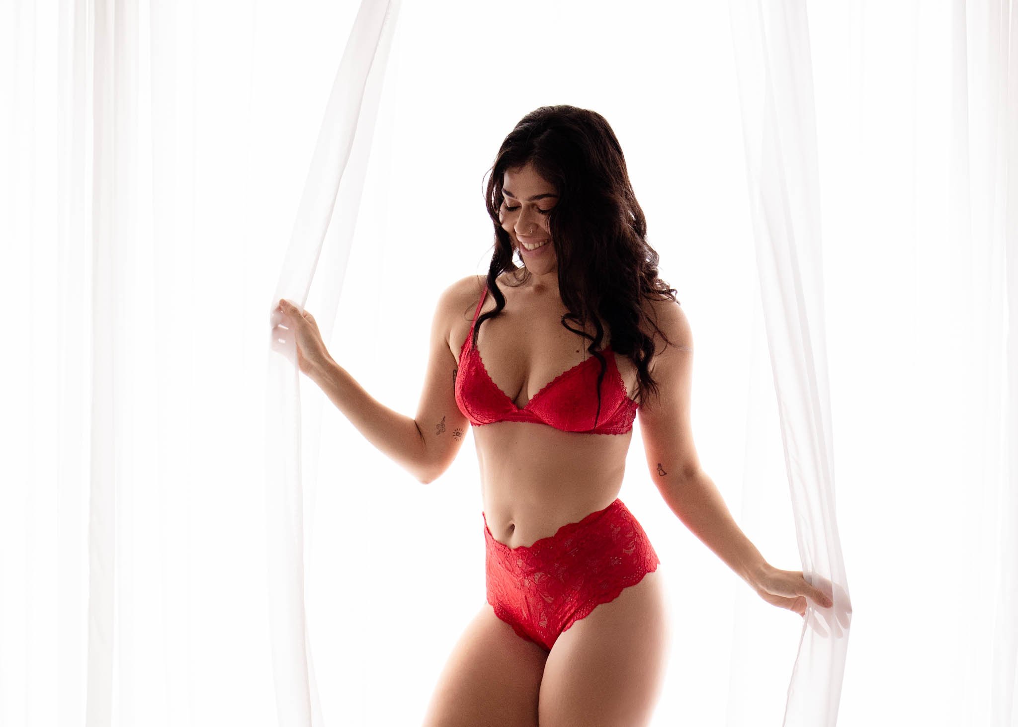 boudoir standing post woman wearing red lingerie next to backlit window with sheer curtains / Photo by Erin Valkner Photography of New Braunfels, Texas. See more at www.erinvalknerphotography.com/blog