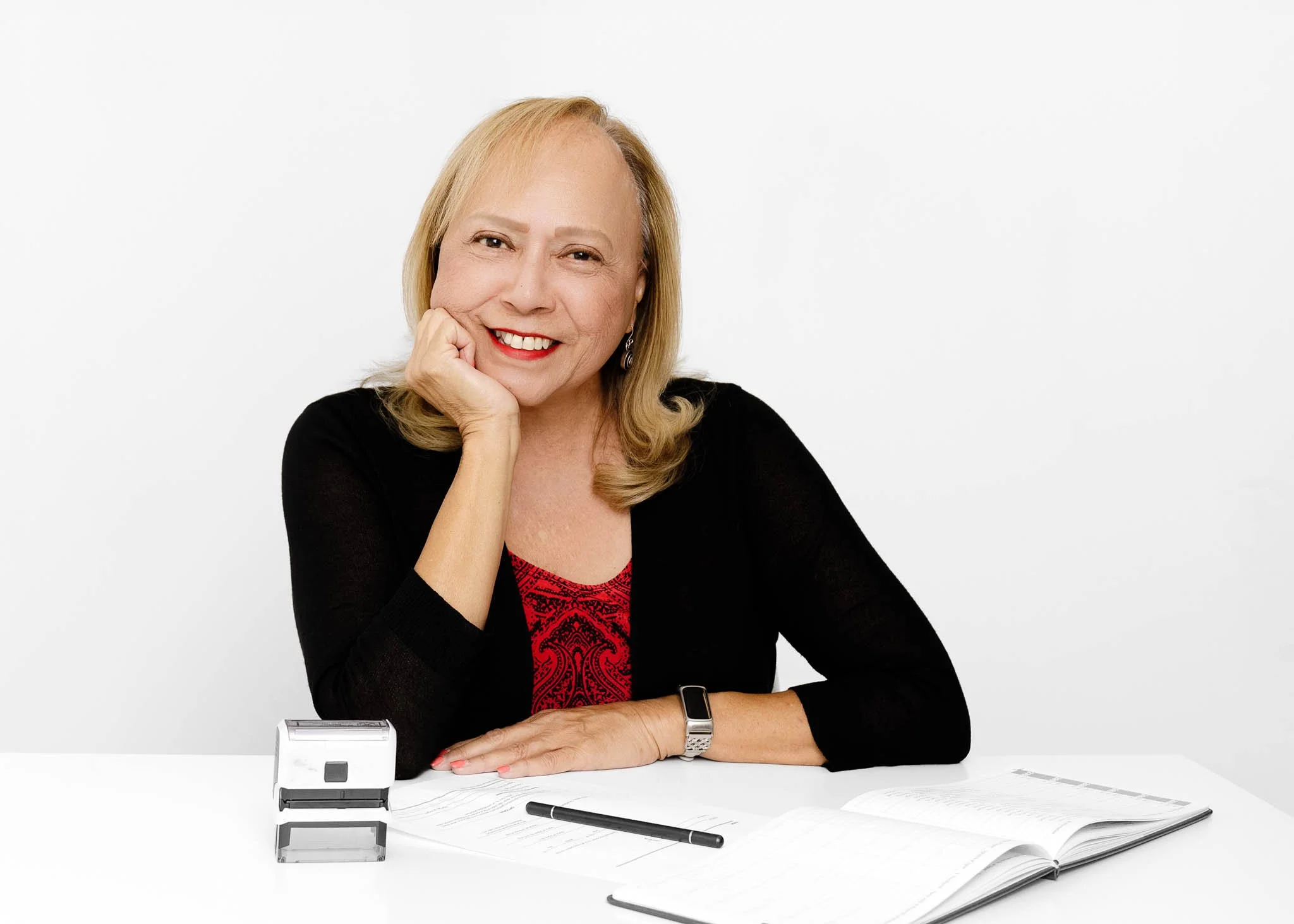 branding photo mature woman sitting at desk relaxed pose smiling looking at camera work wearing red dress and black cardigan blond hair white backdrop