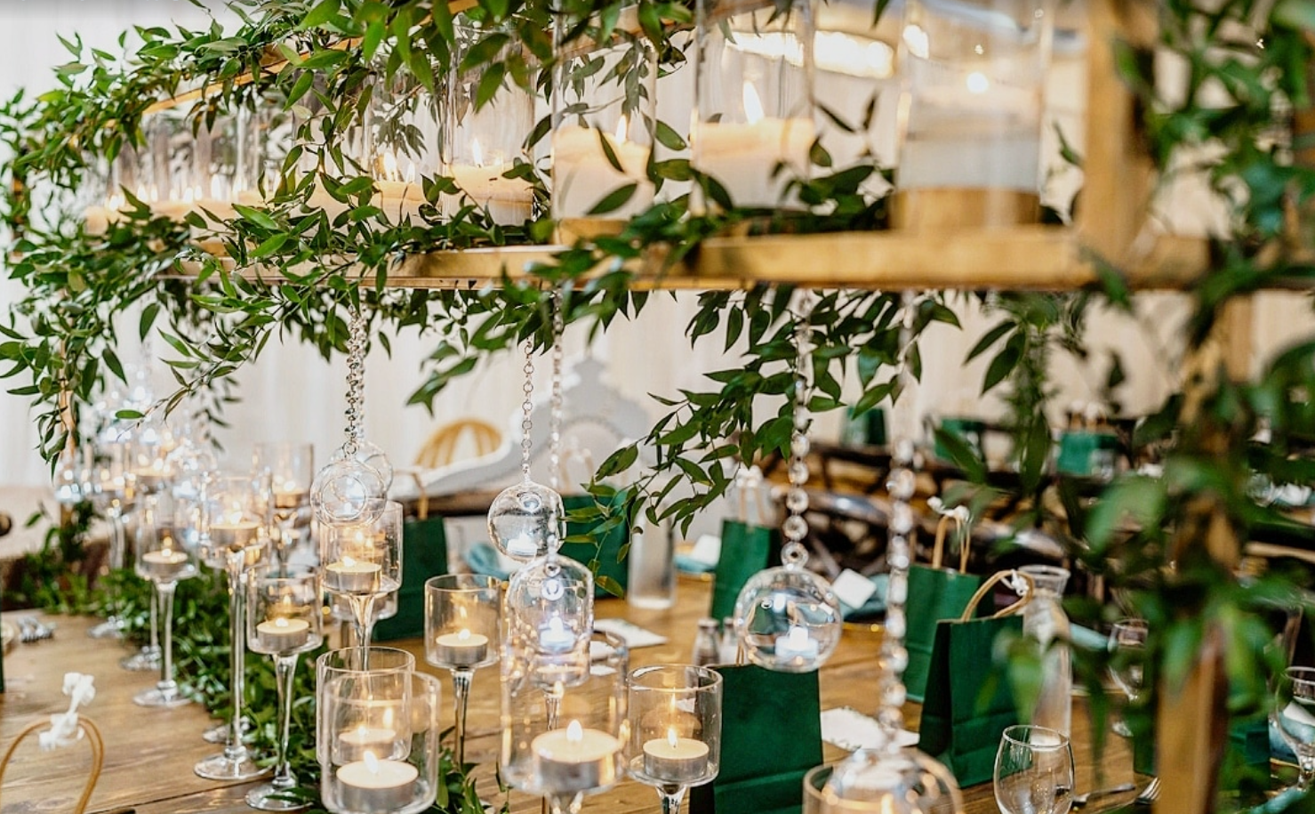 Elegant table setting with candles, glass holders, and green foliage decorations hanging overhead.