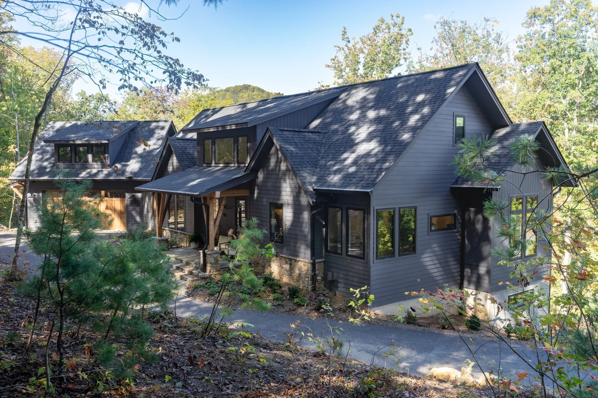 : Exterior of a custom Western North Carolina mountain home with dark gray siding, black-trimmed windows, stone foundation, and timber porch posts surrounded by trees