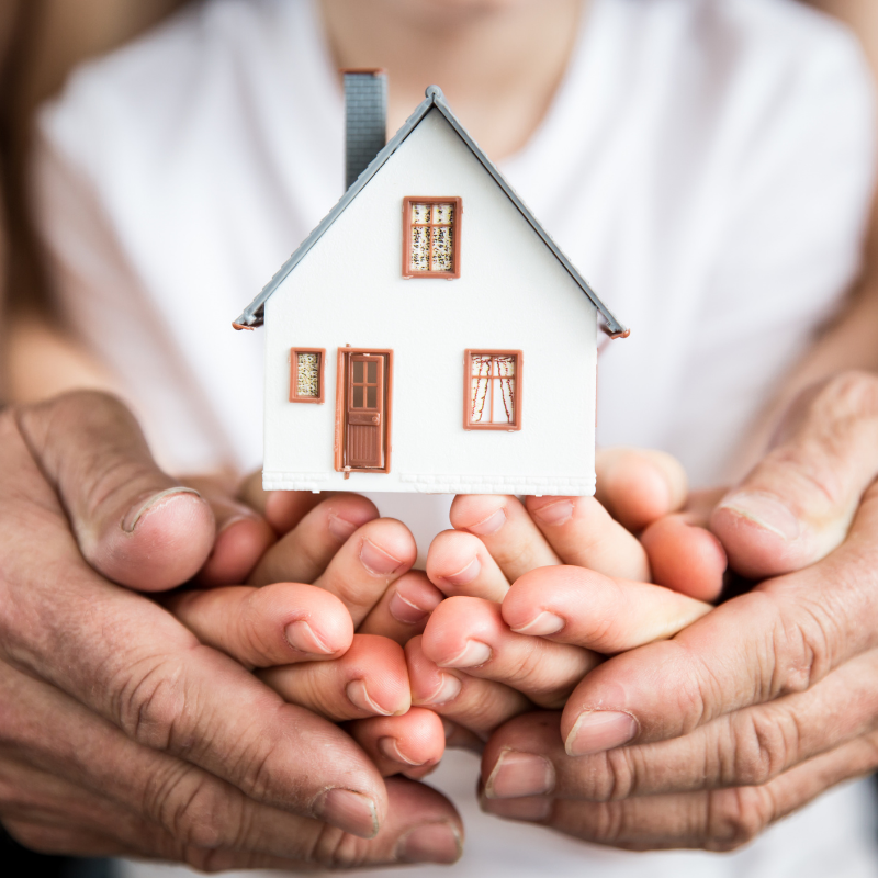 Multiple generations of hands cradling a small model home, representing multigenerational homeownership in Western North Carolina