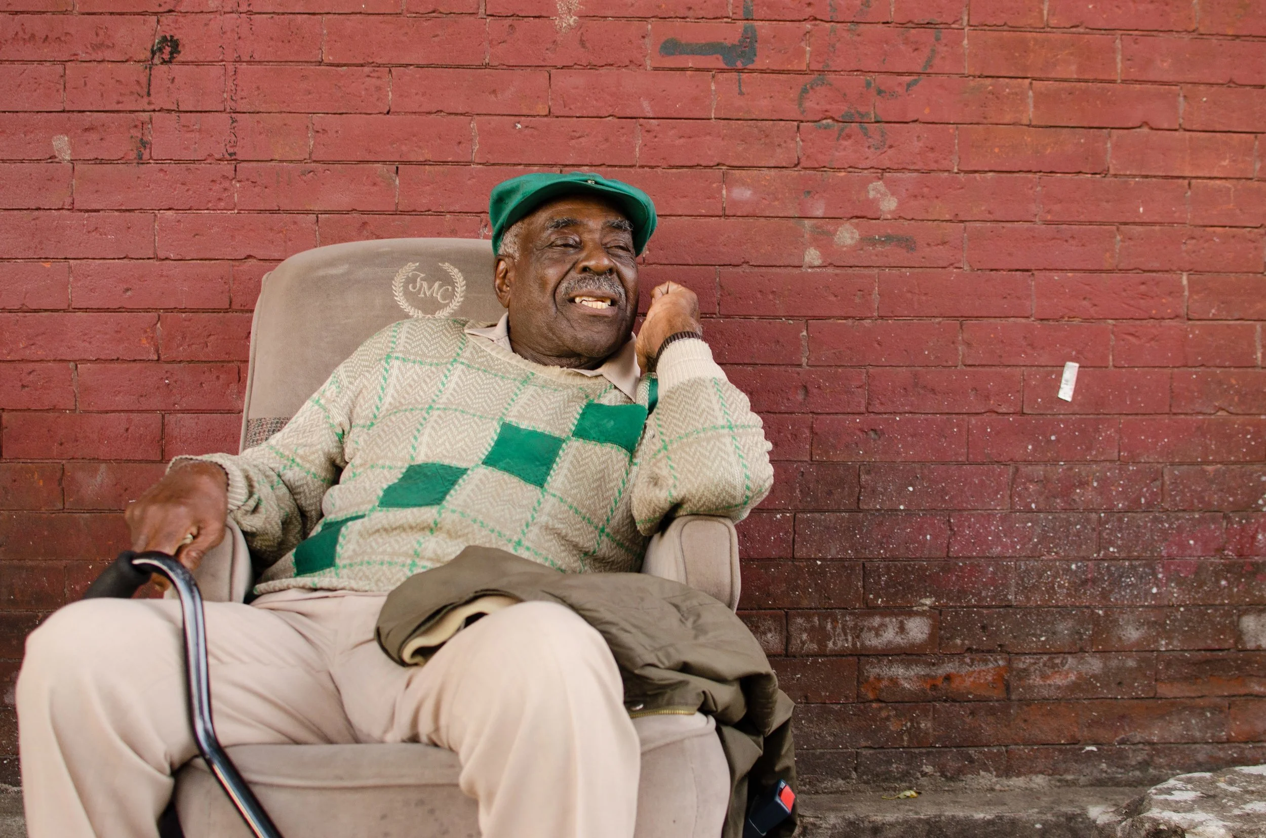 Image: An older Black man reclines in a tan suede chair with his left elbow resting on the chair’s armrest. His metal cane rests on his right leg. Behind him is a red brick wall with markings and imperfections on it. He gazes to the right of the camera with a open-mouthed smile on his face. He is wearing beige slacks, a beige shirt and a green, white, and a beige sweater with a green hat. Photo courtesy of the artist.