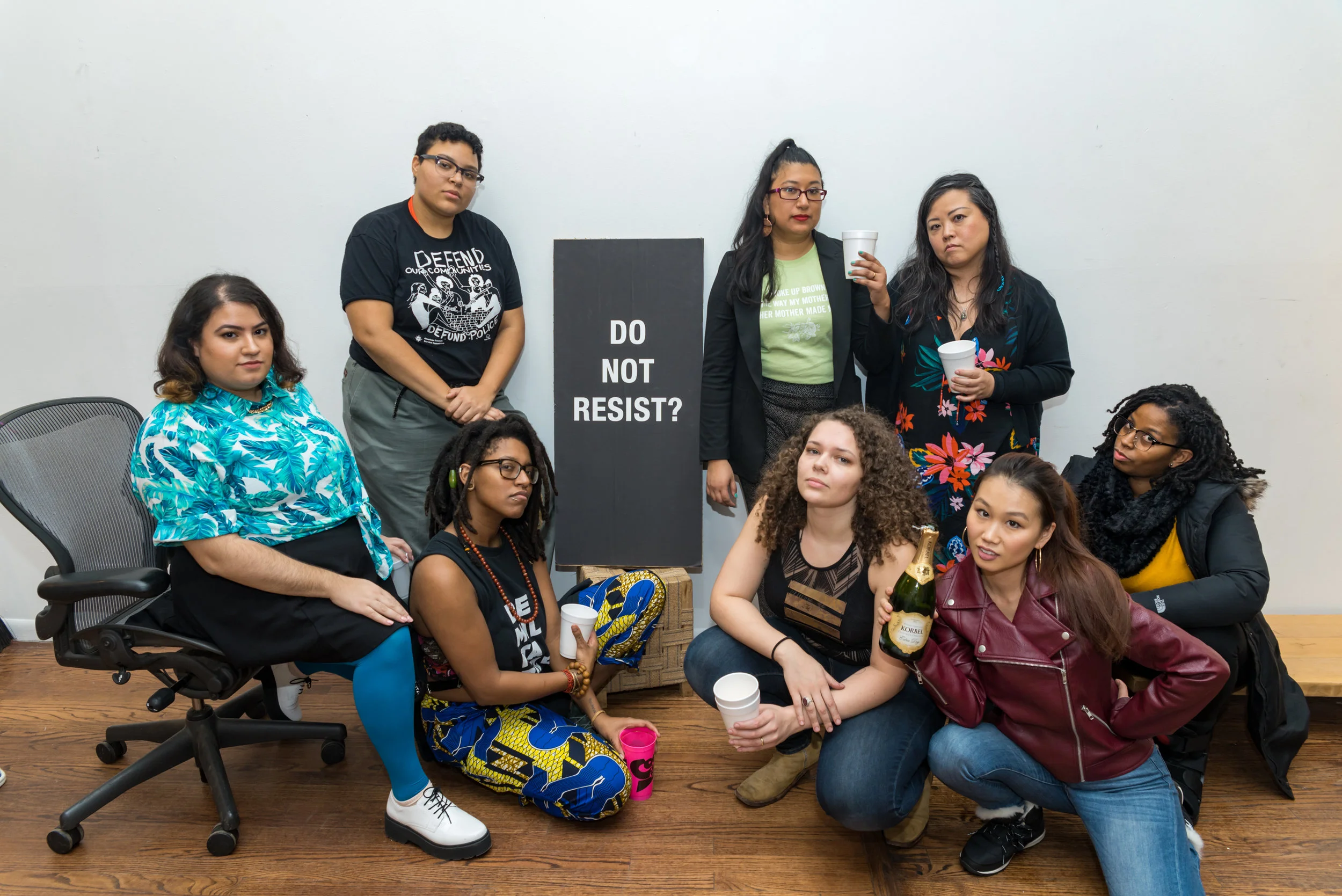 Image: Photograph of eight members of For the People Artists Collective at their Third Annual December Showcase on December 2nd, 2018. They are all looking directly at the camera while posing around a black poster with white coloring that reads “Do Not Resist?”. Photo courtesy of Sarah-Ji of Love and Struggle Photos.