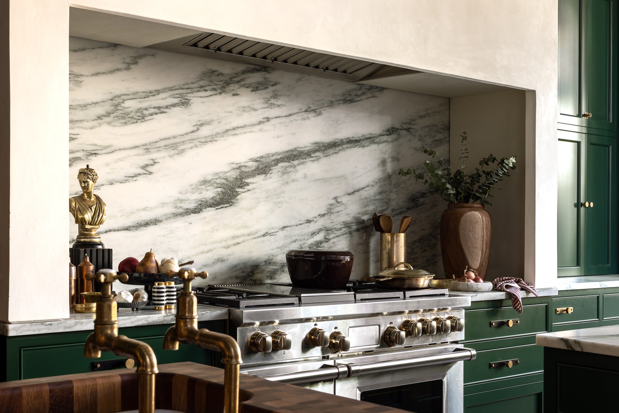 Modern kitchen with green cabinets, marble backsplash, stainless steel stove, brass fixtures, and decorative items including a bust, vase with greenery, and kitchen utensils.