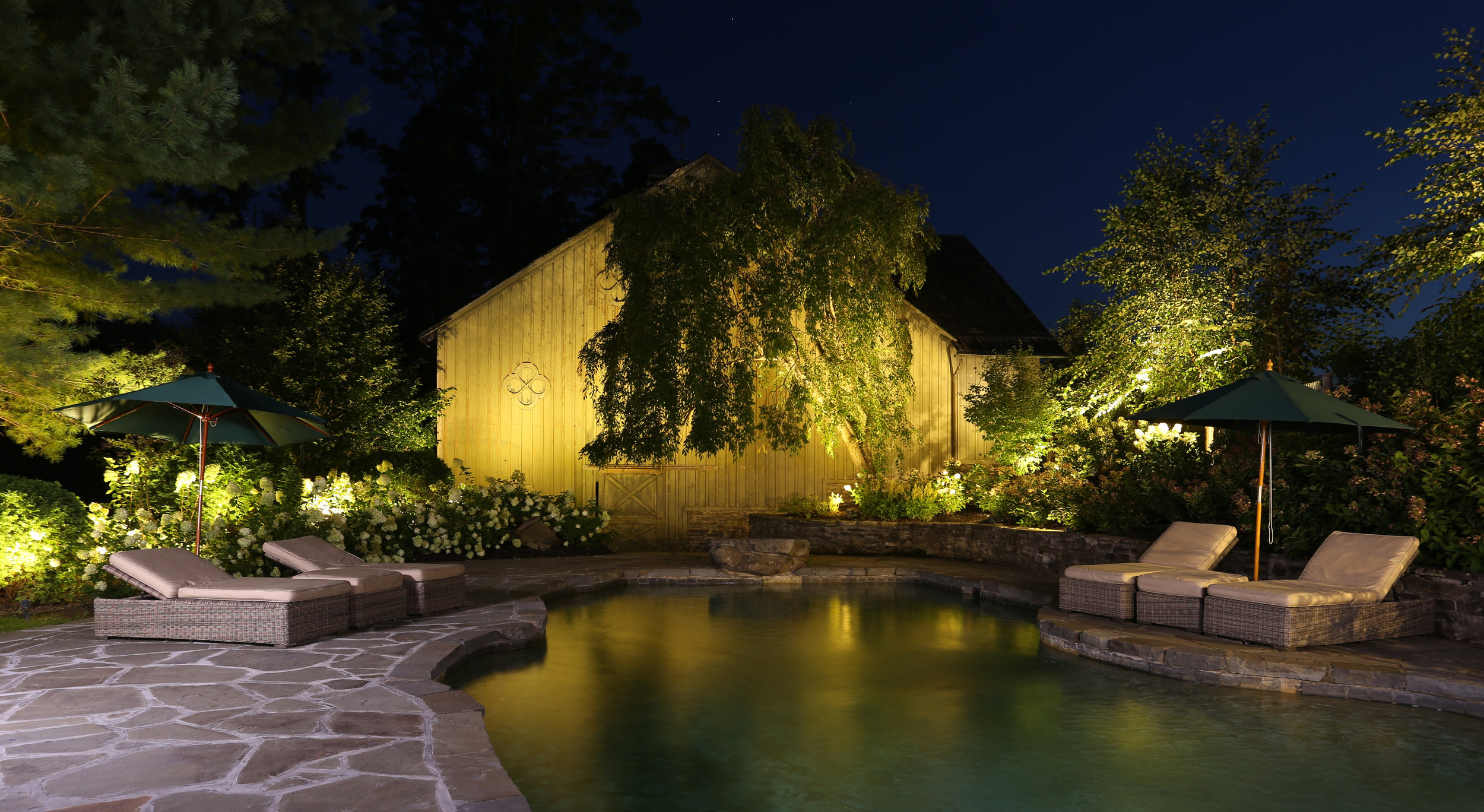 The pool and historic barn glowing at dusk, lit by the estate's landscape lighting