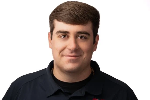 Portrait of a young man with short brown hair wearing a black polo shirt, smiling gently, against a plain white background.