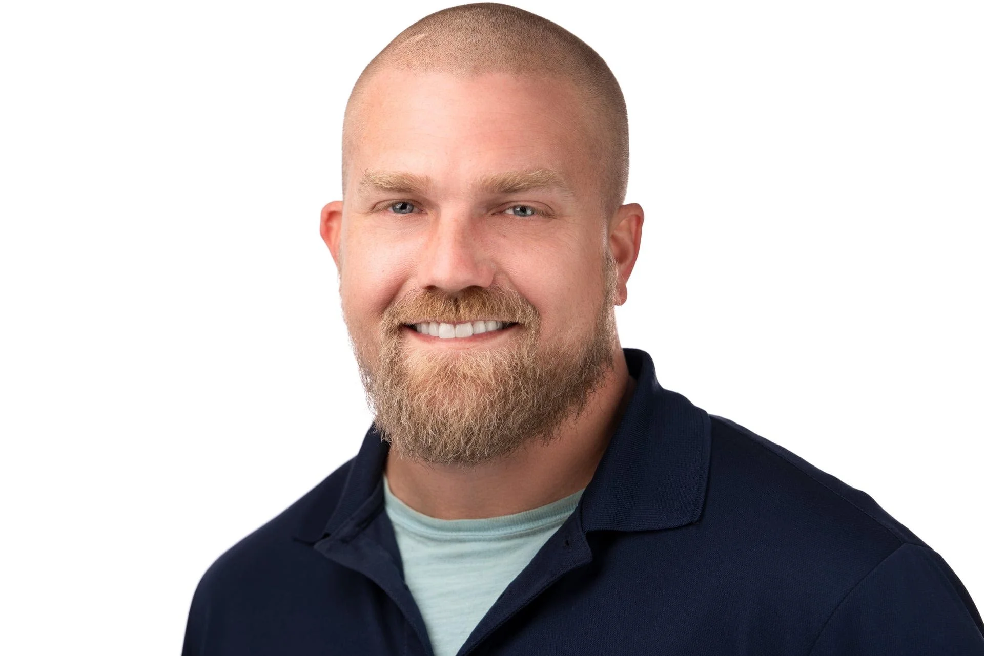 Portrait of a man with a close-cropped haircut, blue eyes, and a beard, smiling and wearing a navy blue shirt against a white background.
