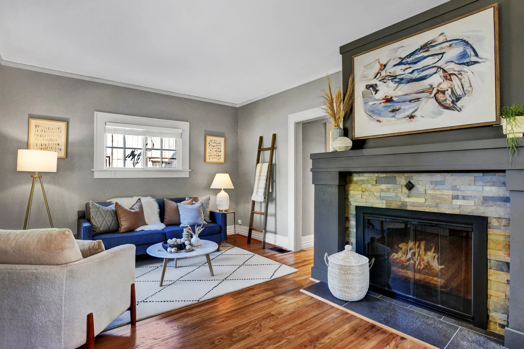 Modern living room with a gray couch, decorative pillows, and a low round coffee table atop a patterned rug. A large abstract painting hangs above a stone fireplace. A floor lamp, window, framed art, and a blanket ladder are also present. Hardwood floors and neutral tones create a cozy atmosphere.