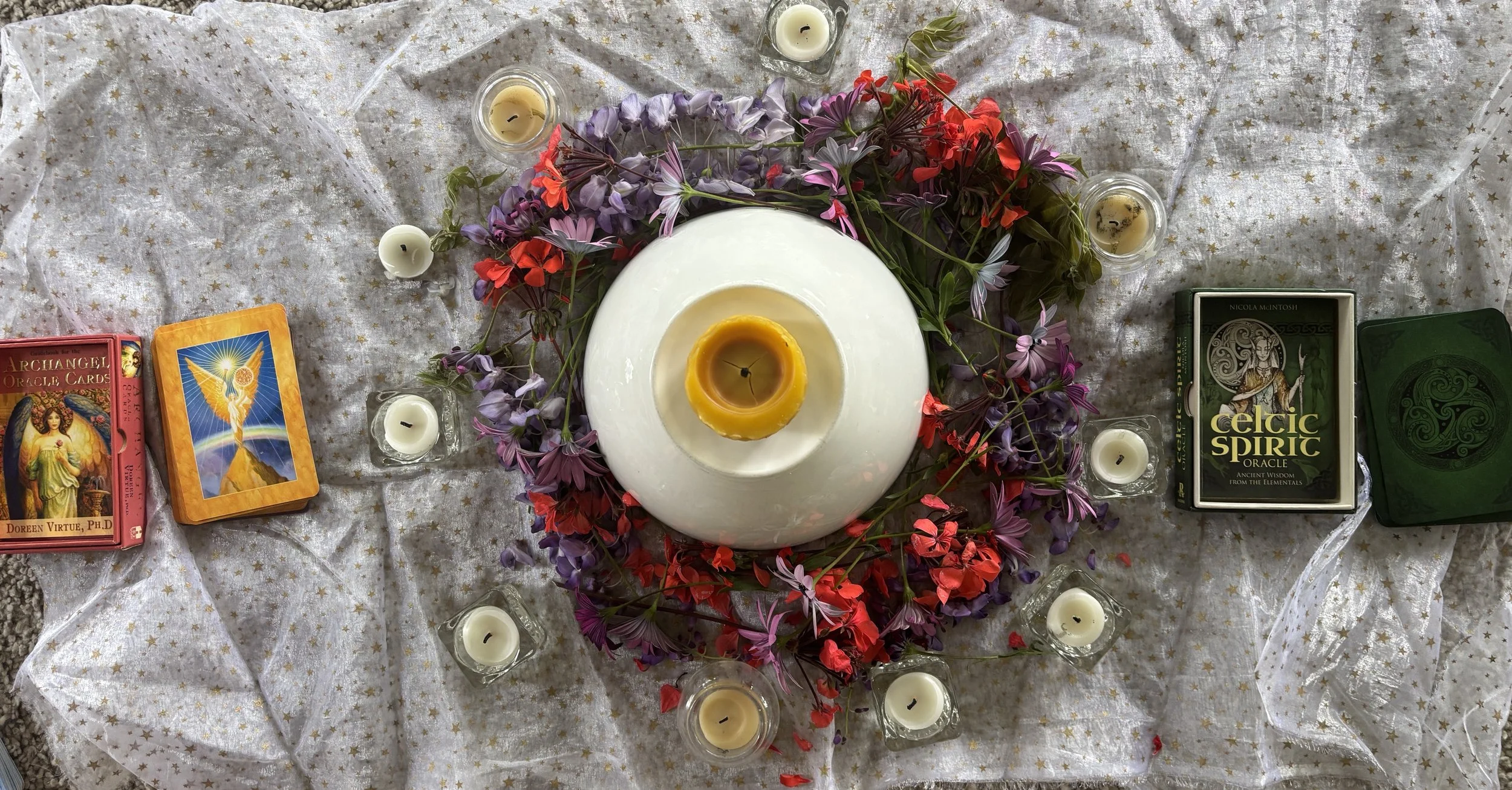 A Mothers' Blessing circle alter setup with a white tablecloth and gold star pattern, surrounded by candles and colorful flowers, with angel and divine feminine cards on either side. beads, red thread, crystals,