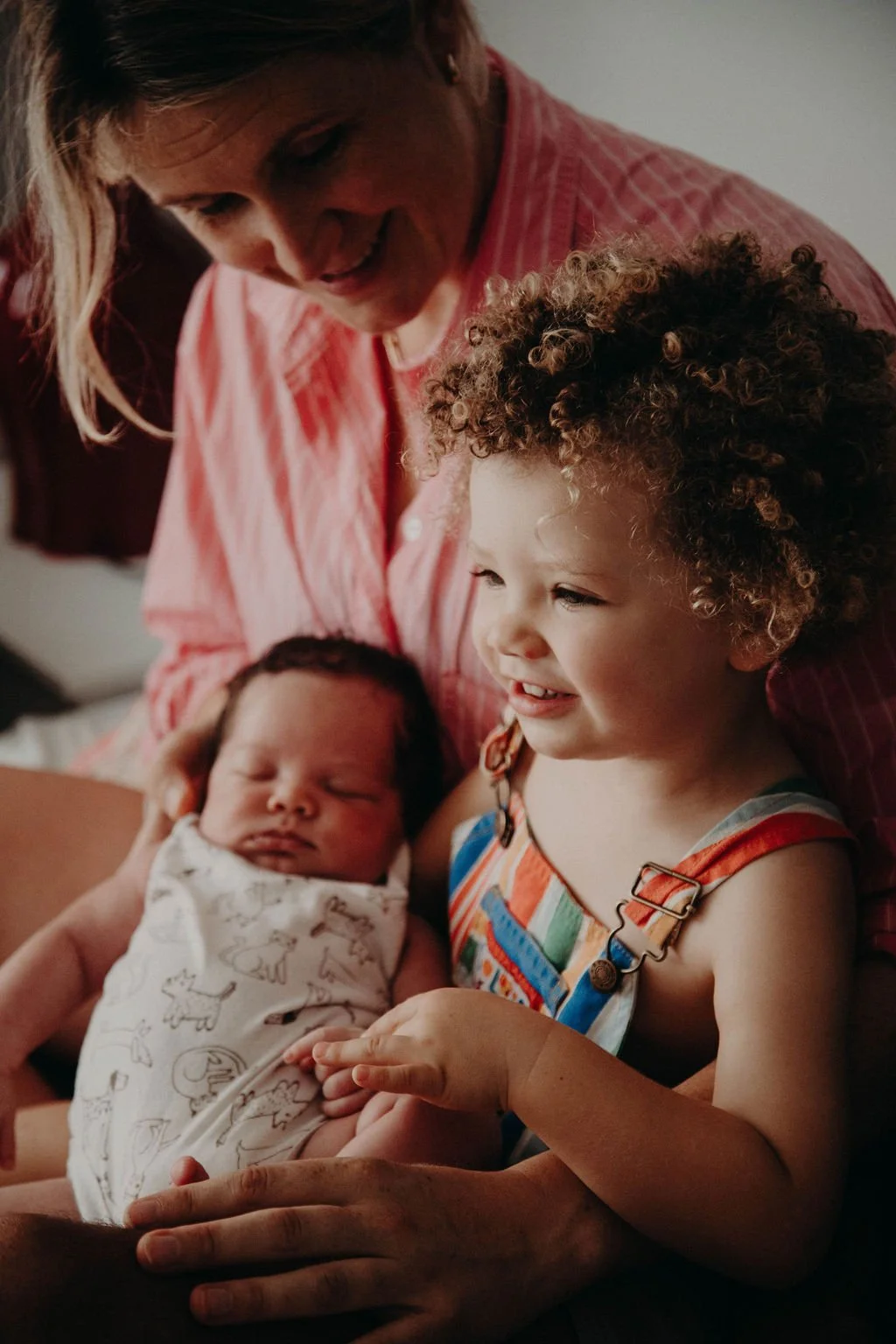 A woman with two children, one a newborn and the other a young child, holding and showing the children to each other in a warm, intimate moment. Postpartum doula one-off care individual session from mothers with babies up to 12 months.