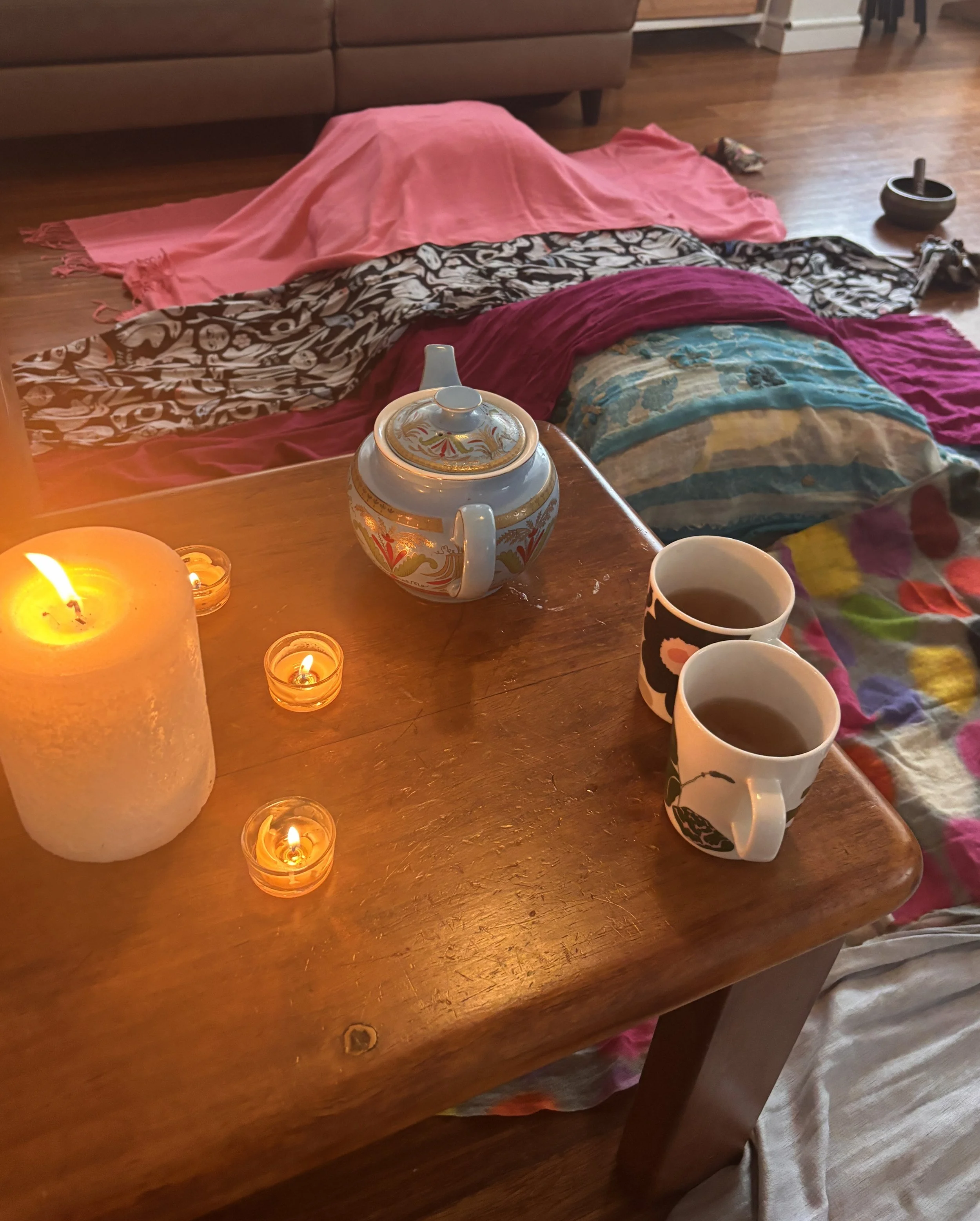 A cozy indoor scene with a wooden table illuminated by candles, including a large candle, smaller tea light candles, and a ceramic teapot with floral and gold accents. A mug filled with a hot beverage and colorful cloths or blankets are visible. The background features a brown couch and hardwood flooring.