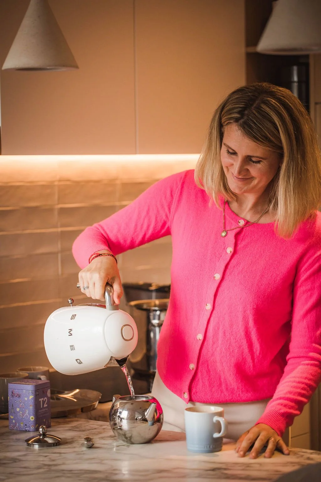 A woman in a pink sweater pouring hot water from a kettle into a teapot in a home kitchen, reflecting gentle, practical postpartum care and nourishment.