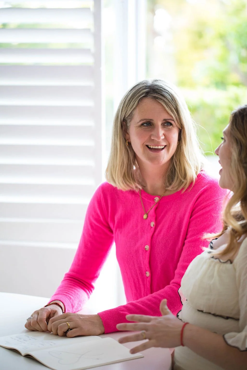Two women sitting at a table, engaged in conversation indoors near window with closed shutters, one woman smiling, the other gesturing with hands.