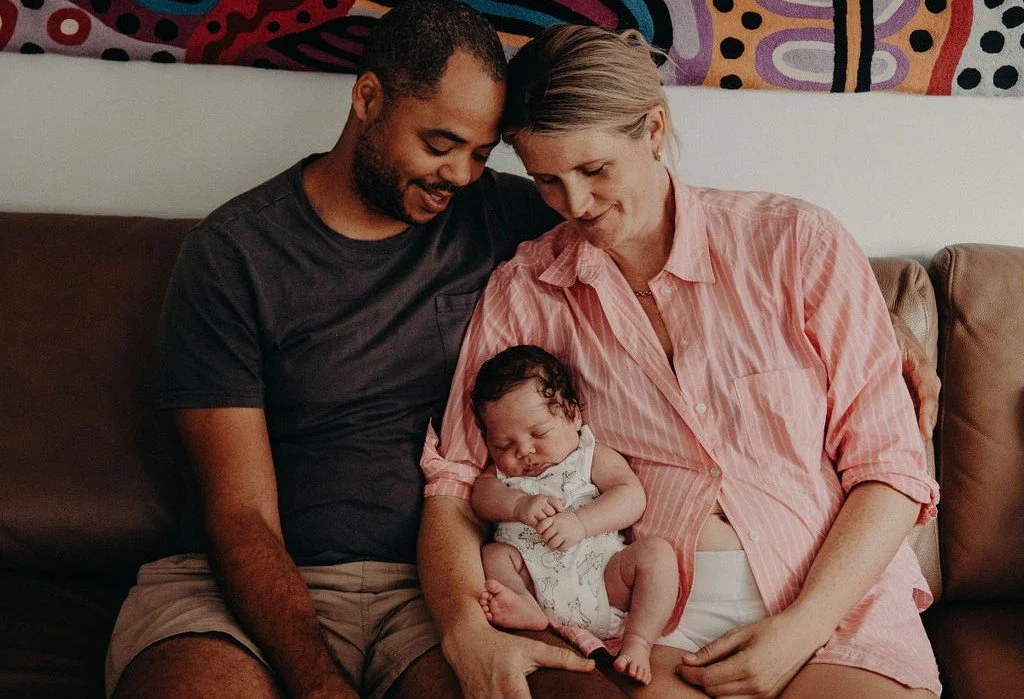 New mum and dad look down at newborn baby, while seated on a brown couch