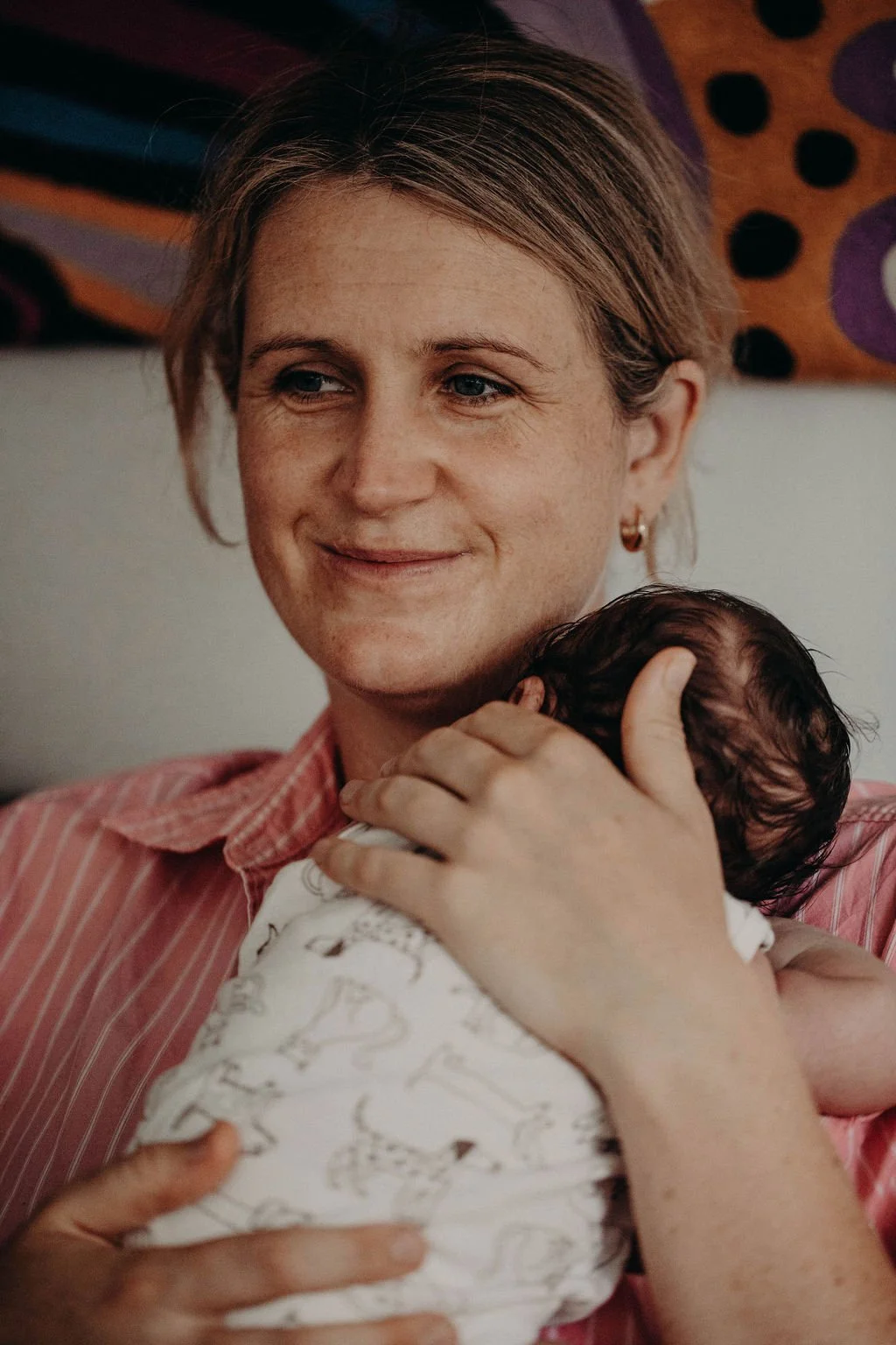 A woman with light brown hair, wearing small hoop earrings and a pink striped shirt, holds a newborn baby with dark hair closely against her chest, smiling softly. What we might do during a Postpartum Doula home visit. You rest and I care for you.