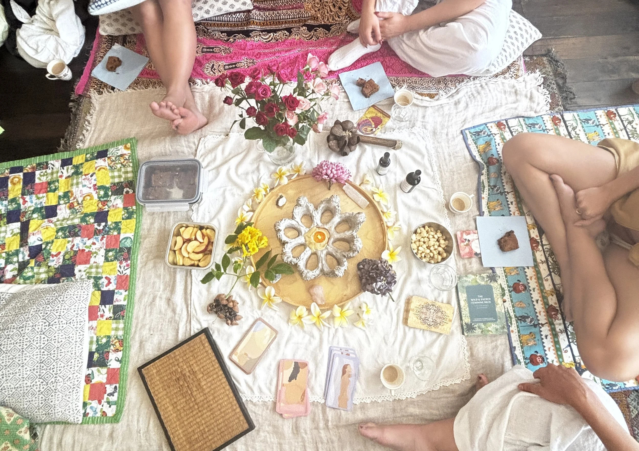 People sitting around a table with a floral centerpiece, snacks, and tarot or oracle cards, engaging in a spiritual or social gathering.