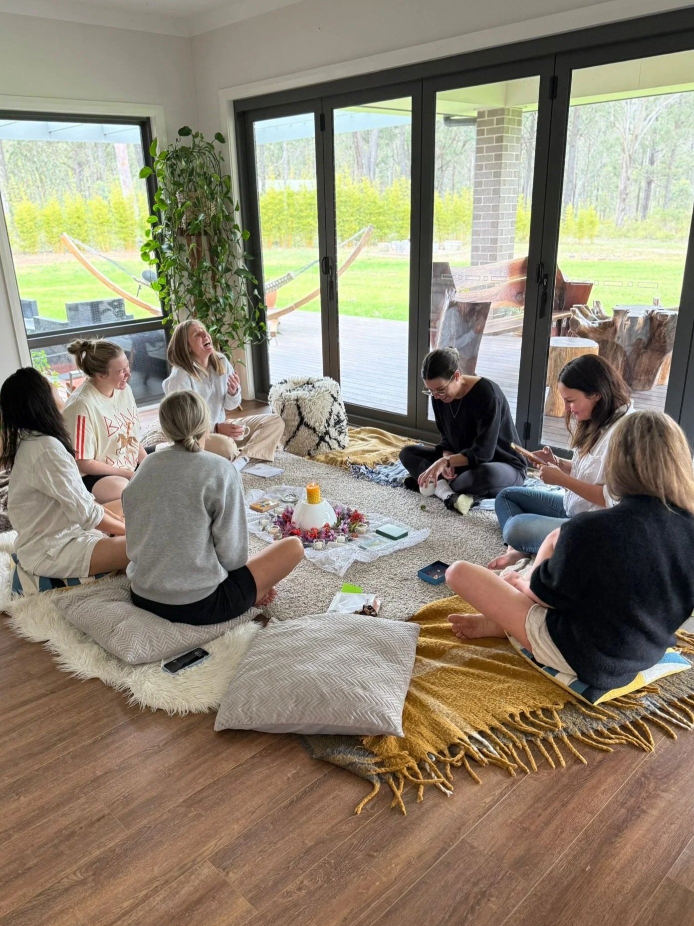 A group of nine people sitting at a Mothers' Blessing on cushions and a rug on the wooden floor inside a room with large windows, sharing laughs, with a small table in the center holding candles and decorations, facilitated by Hats from Pink Flamingo