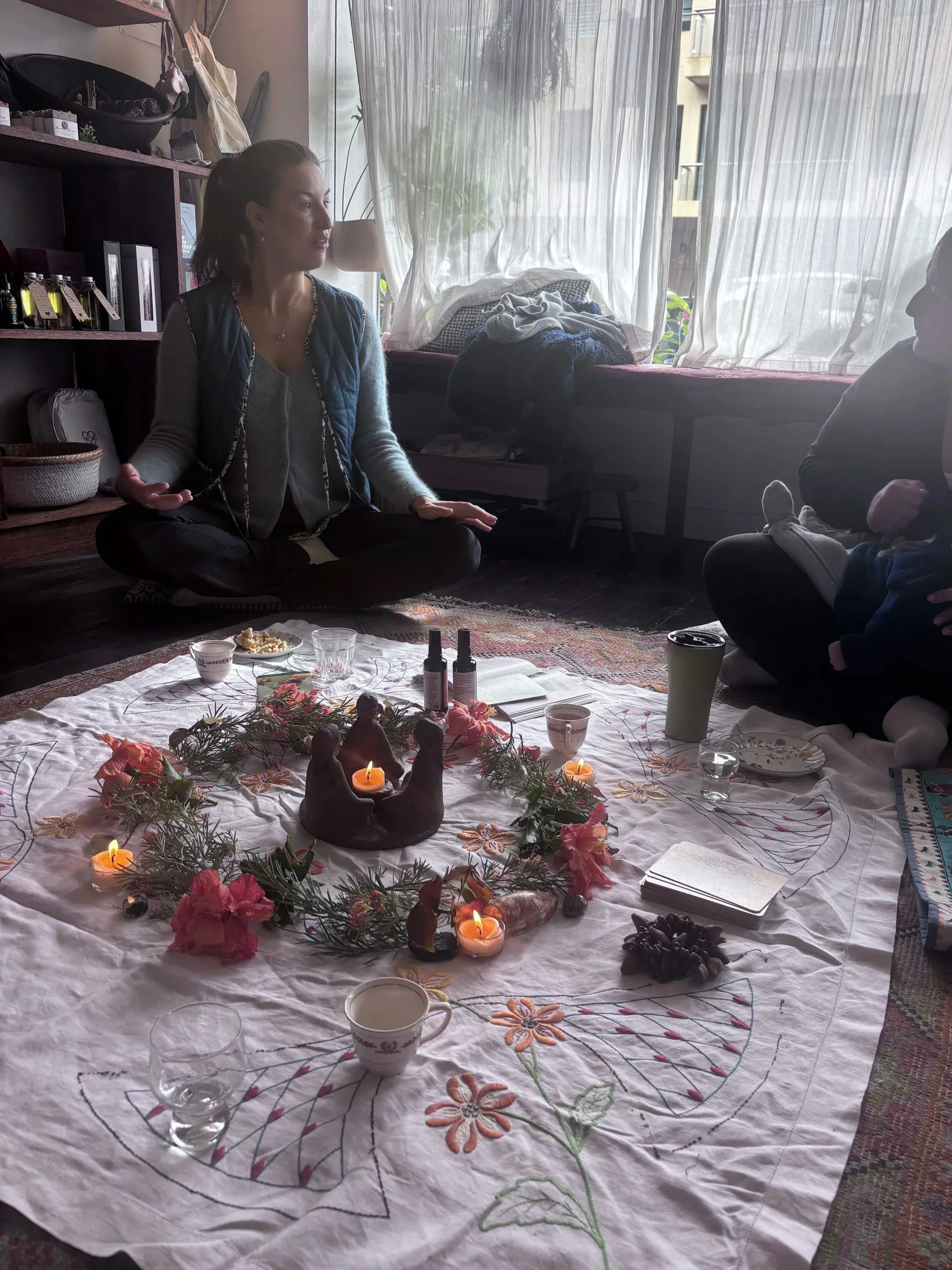 Indoor scene of a Mothers' Circle with a large decorated tablecloth, adorned with candles, flowers, and crystals. A pregnant and a postpartum woman are sitting on the floor, engaged in a conversation near a window with sheer curtains. Eastern Sydney