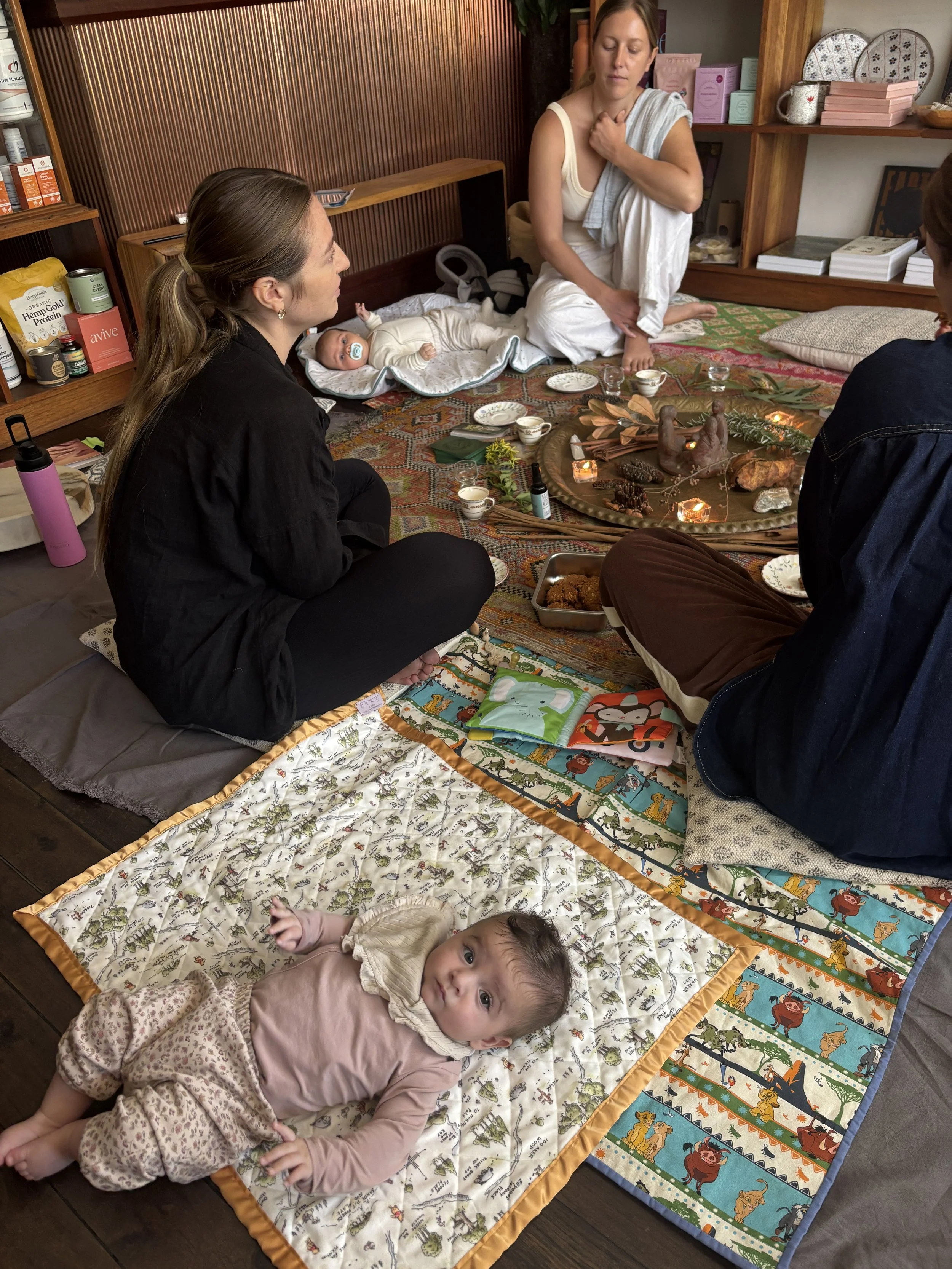 A group of people and babies sitting on colorful blankets on the floor in a cozy room, with a decorated tray of candles and natural objects in the center, possibly during a meditation or mindfulness session.