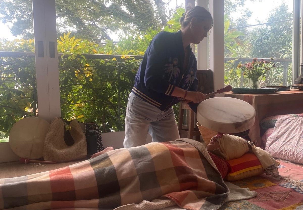 Hats uses a shamanic drum and hands-on touch in a womb healing or Closing of the Bones ritual to bathe a person lying, resting, relaxing on the floor in beautiful vibrations,The room has large windows and green plants outside.