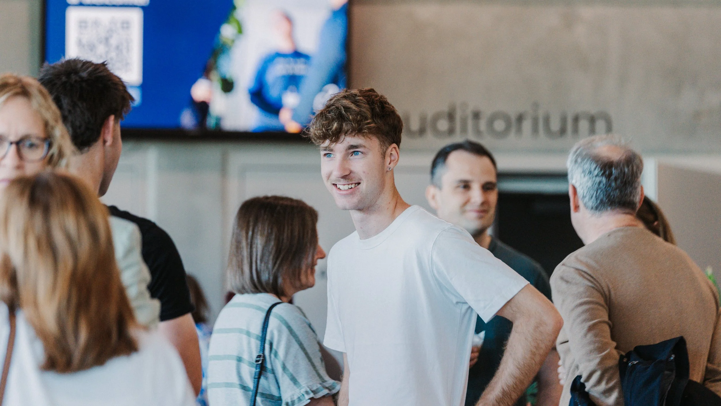 A group of people socializing indoors, with a young man smiling at the center, surrounded by others engaged in conversation.