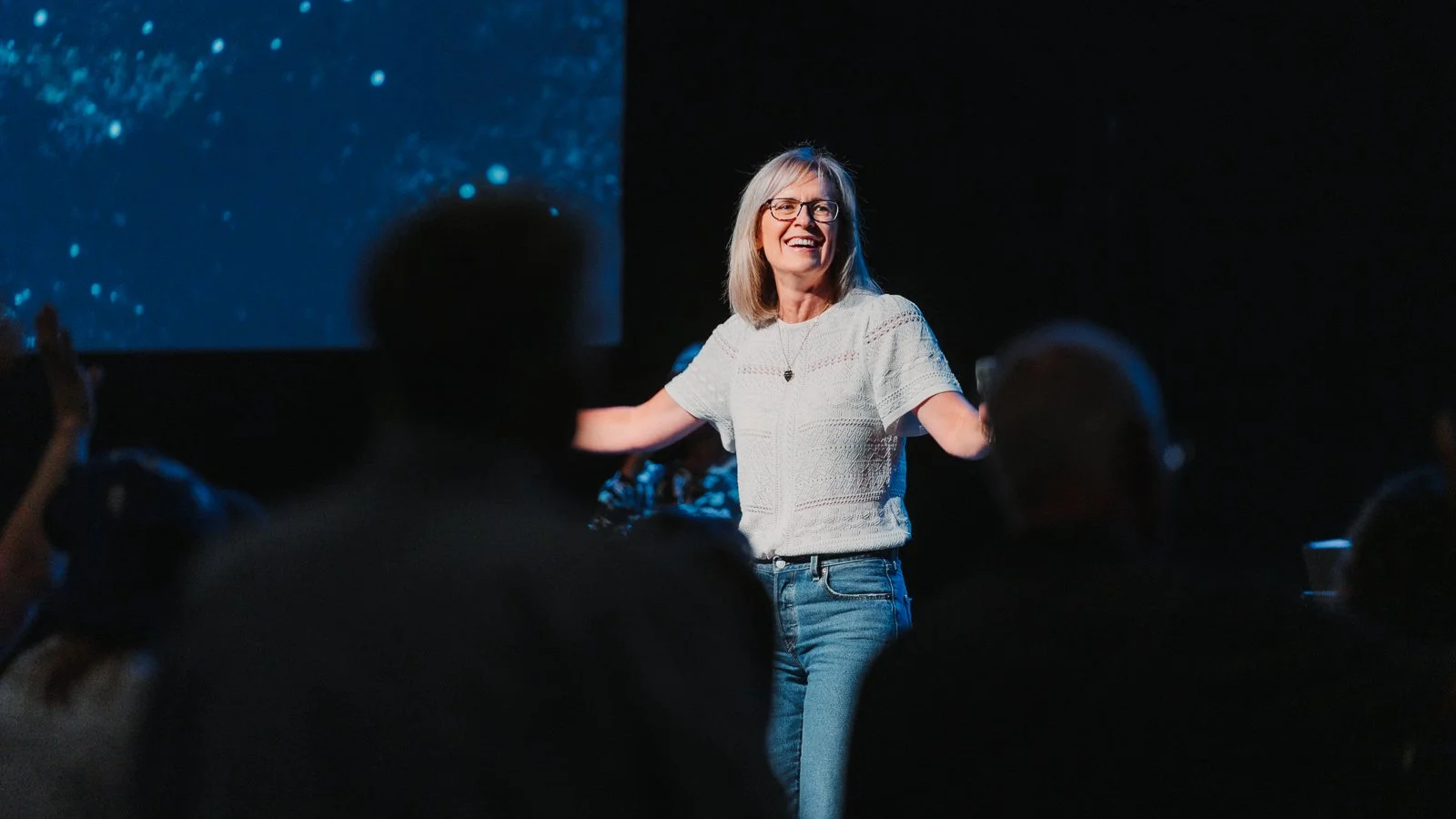 A woman on stage smiling with open arms during a presentation, audience members in the foreground, dark background with a starry sky projection.
