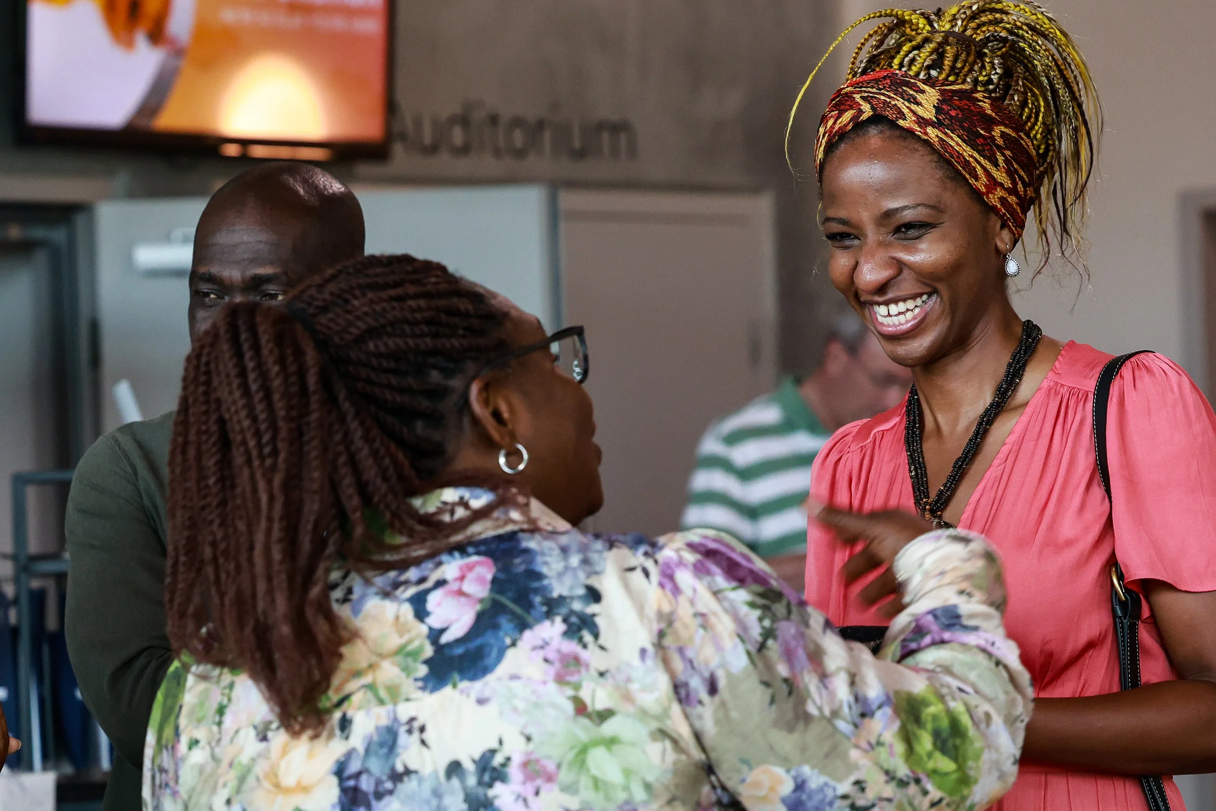 A woman with yellow and black braided hair and a colorful headwrap, smiling and talking to an older woman with glasses and braided hair, in an indoor setting.