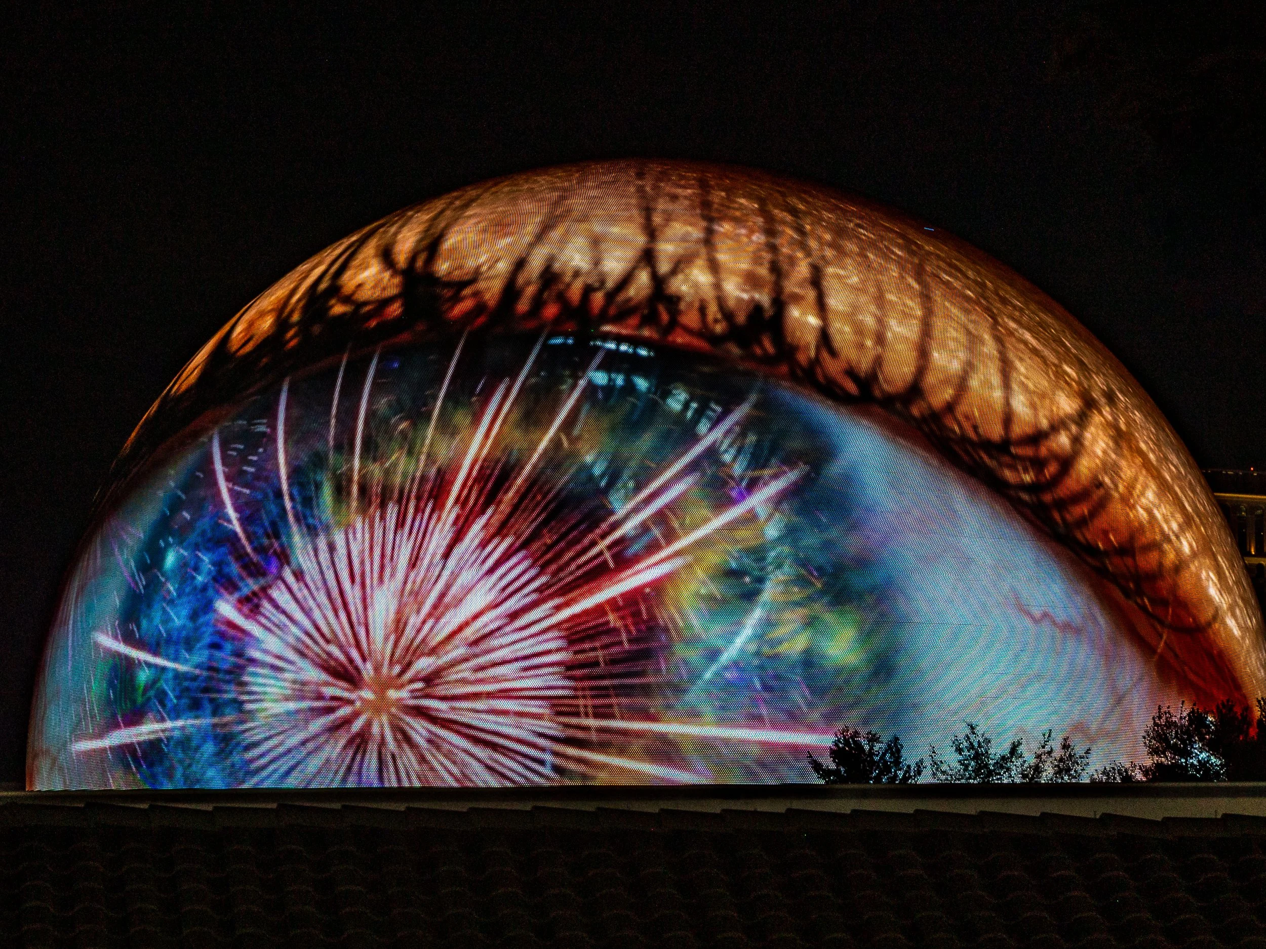 A large illuminated eye-shaped projection display at night, showing fireworks and a tree silhouette.