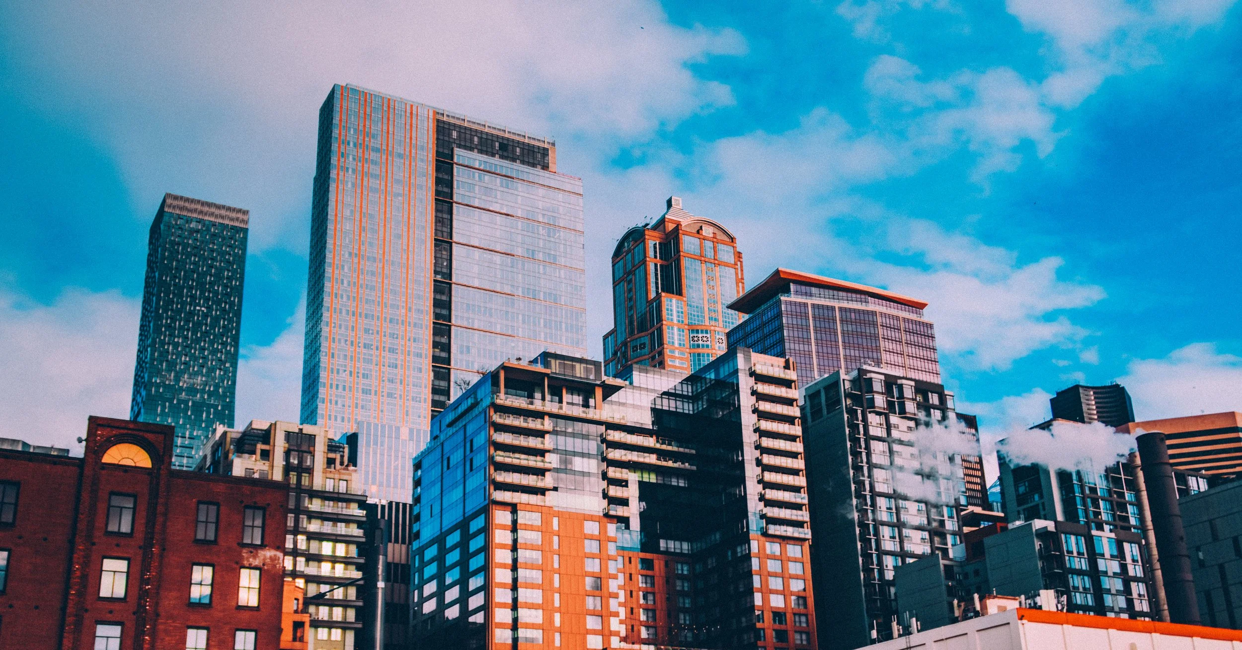 Downtown Seattle city skyline with modern high-rise buildings under a partly cloudy blue sky, some buildings emitting steam from their chimneys.