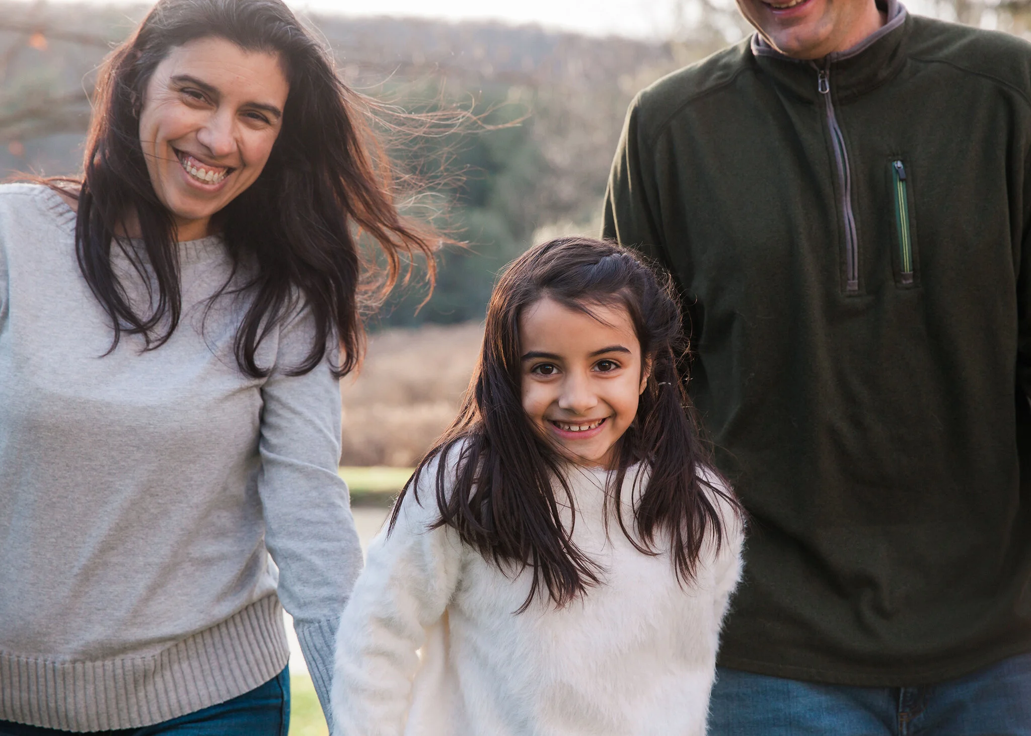 Two parents and a young girl walk toward the camera. The sun is behind them. Hair is blowing in the breeze and fall colors can be seen behind them.