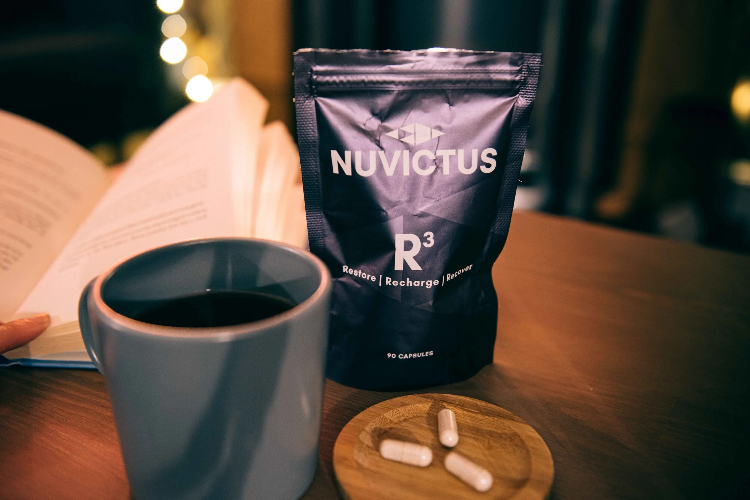A white mug filled with dark coffee, a small wooden plate with three capsules, an open book, and a bag labeled 'Nuvictus R3' with the words 'Restore | Recharge | Recover' on it sitting on a wooden table.