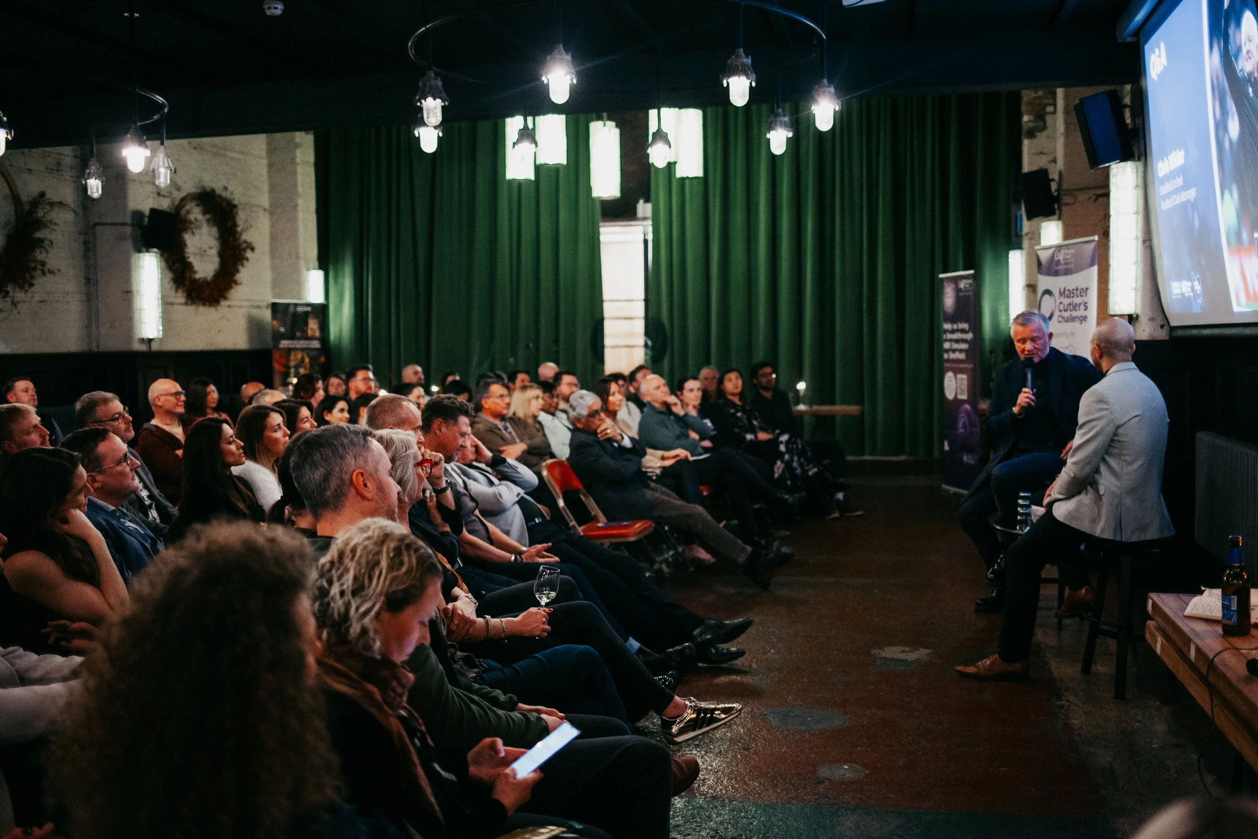 A speaker and an interviewer seated on stage in front of a seated audience in a dimly lit room with green curtains and hanging lights.