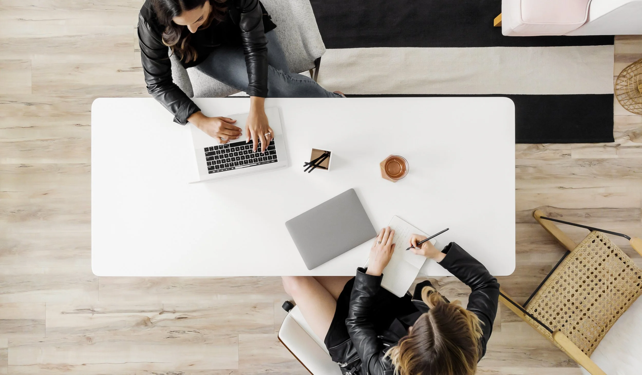 An overhead view of two women working at a white desk with a laptop, notebook, pens, and a cup, in a modern workspace with wooden flooring and minimalist decor.