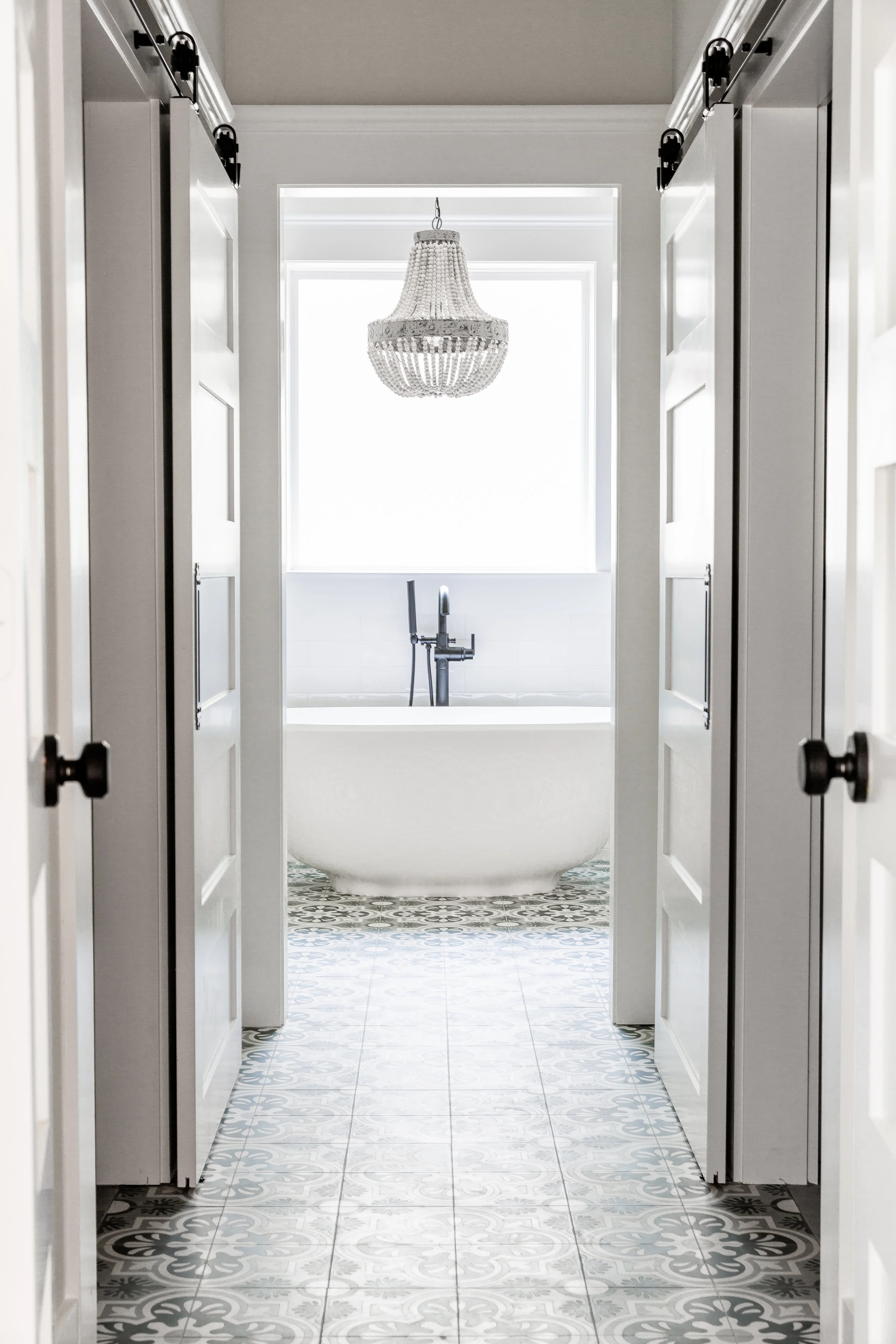 A hallway flanked by barn doors opening up to his & hers closets connects the master bathroom to the existing master bedroom. The bathtub is visible from the entry, anchored under a beaded chandelier by Shades of Light .
