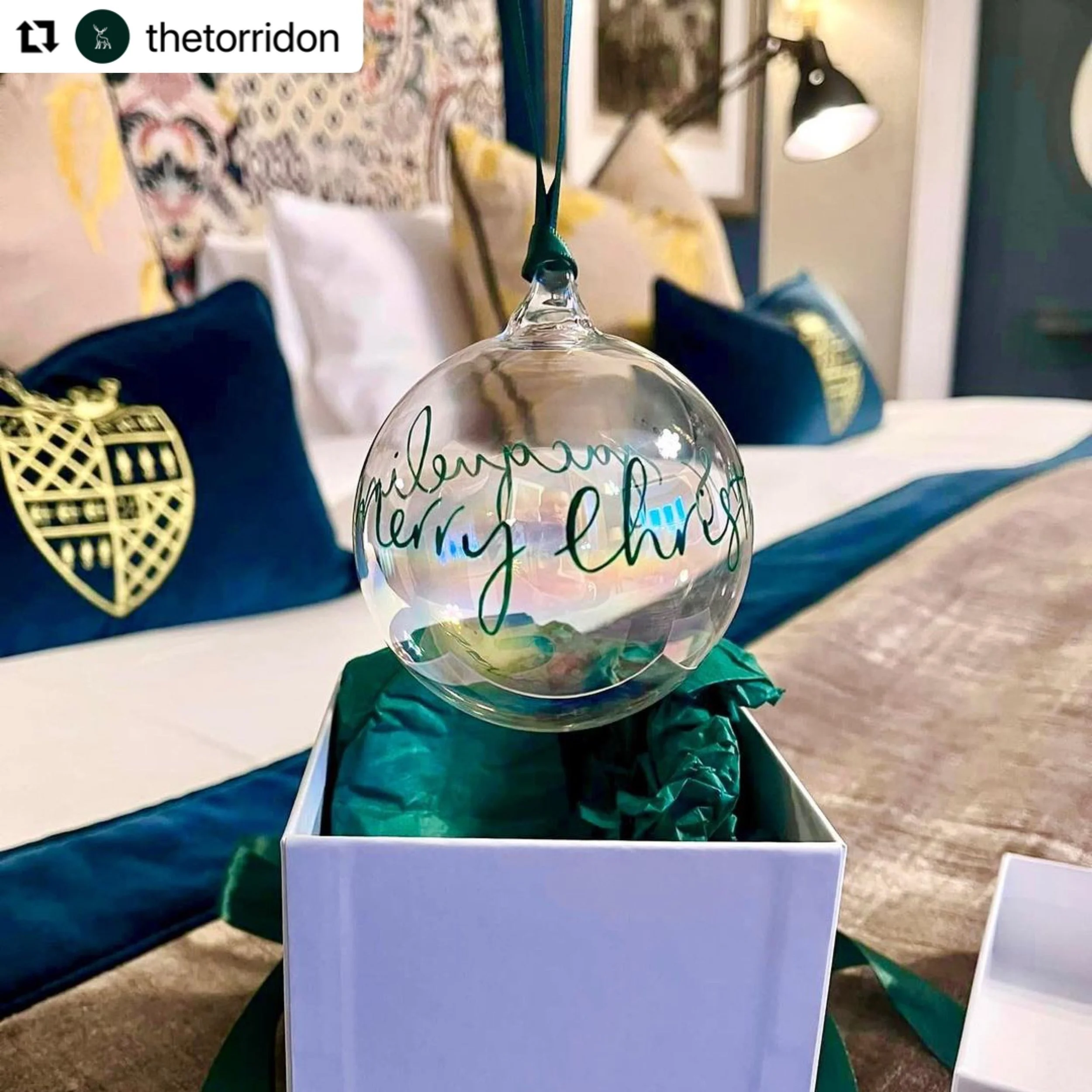 A personalised bauble being dangled above the packaging. In the background is a  guest's bedroom in The Torridon.