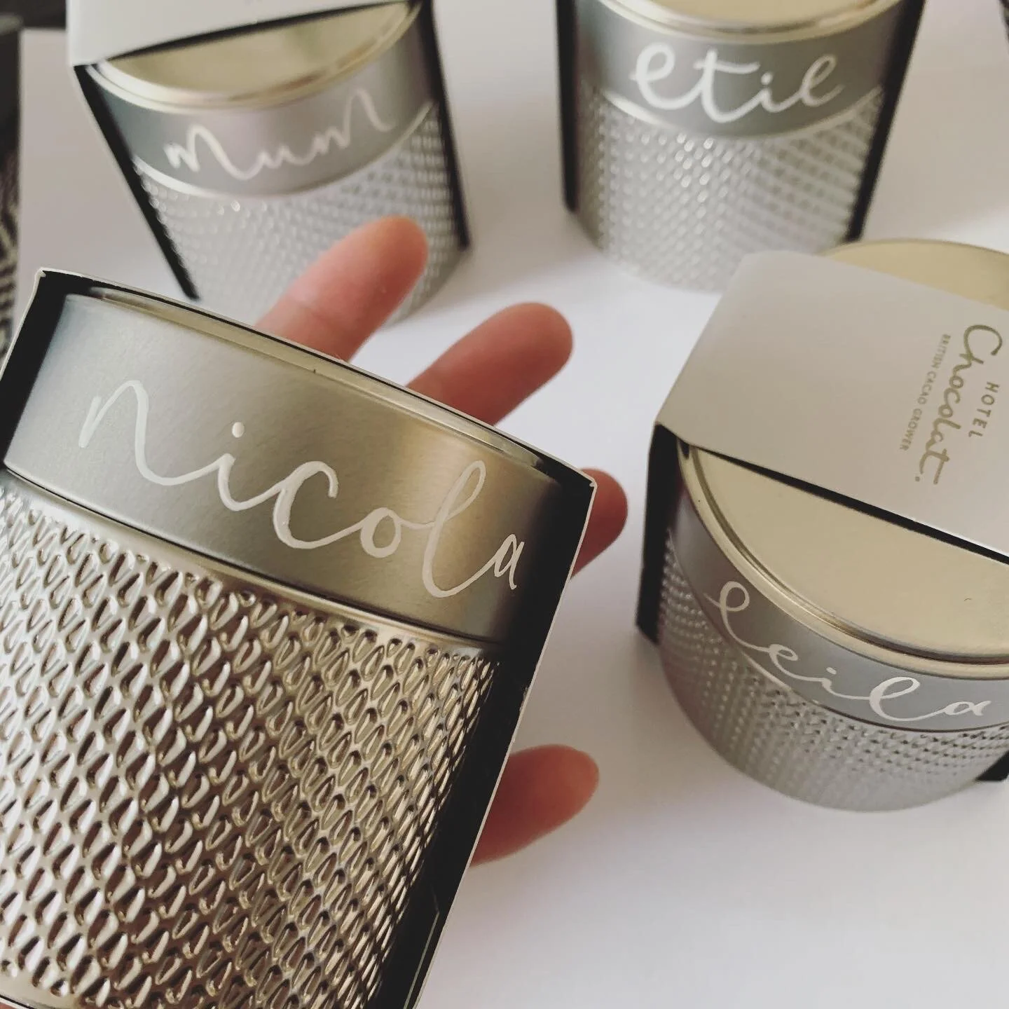Hotel Chocolat tins personalised with guest names in white enamel. A hand is holding a tin with the name 'Nicola'.