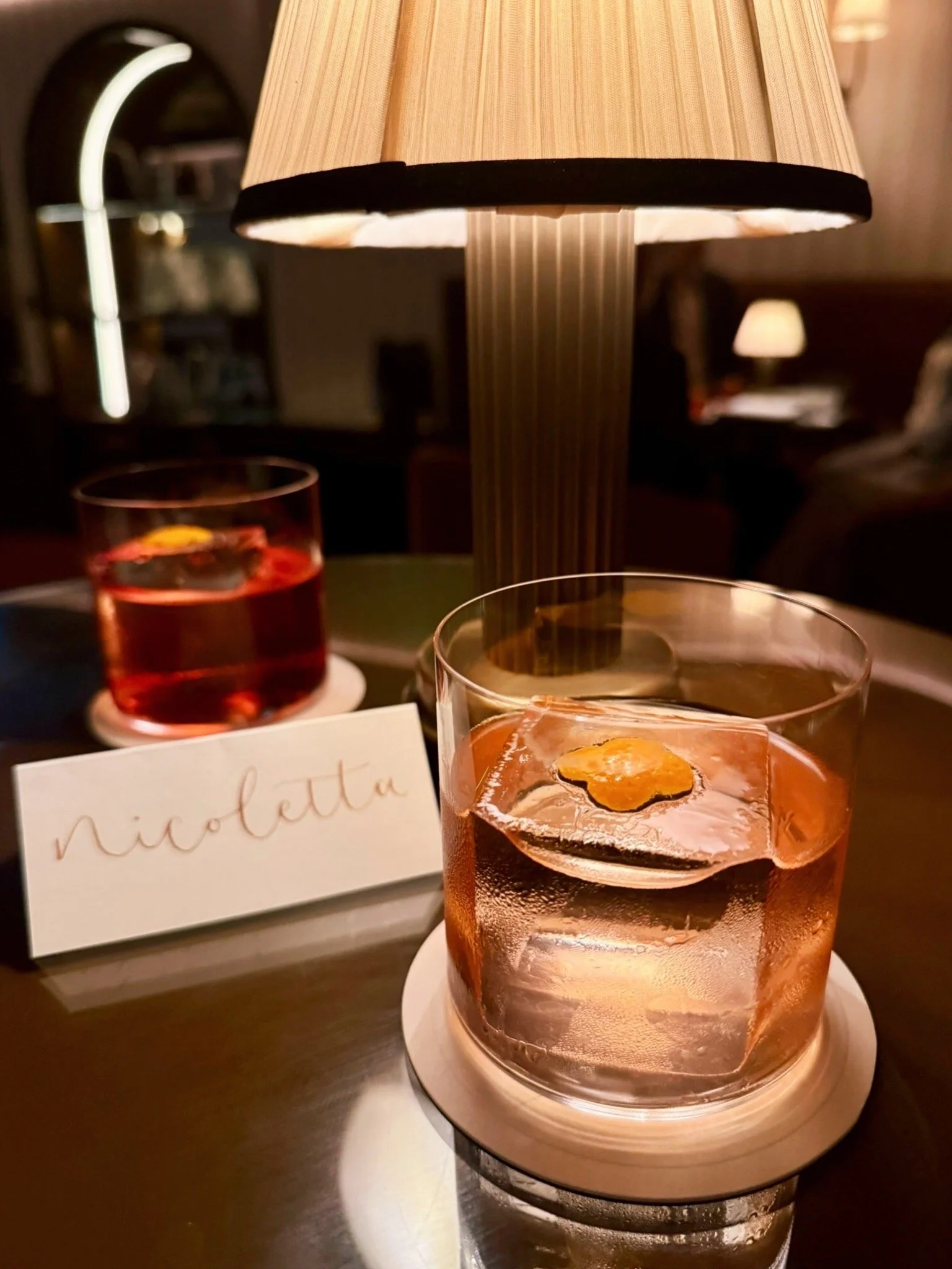 Two cocktails sat next to each other in a posh bar setting, with a place card that has the name 'nicoletta' on it in gold pigment.