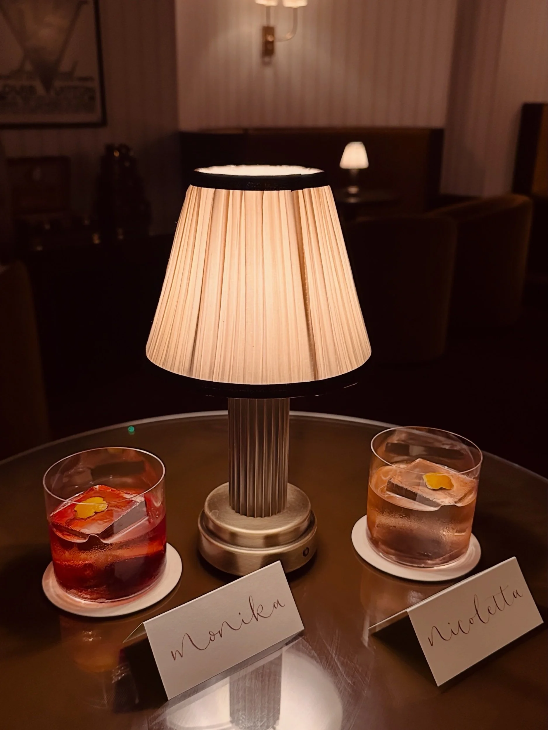 Two cocktails sat next to each other in a posh bar setting. There is a small table lamp behind and two place cards with the names 'nicoletta' and 'monika'.