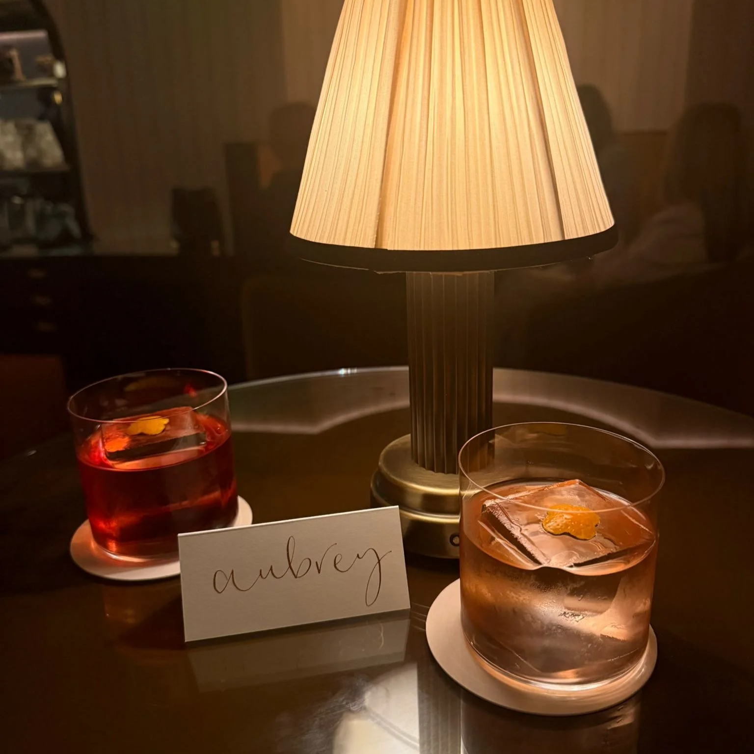 Ivory place card that displays the name 'aubrey' in gold pigment, sat next to two cocktail glasses on a little bar table.