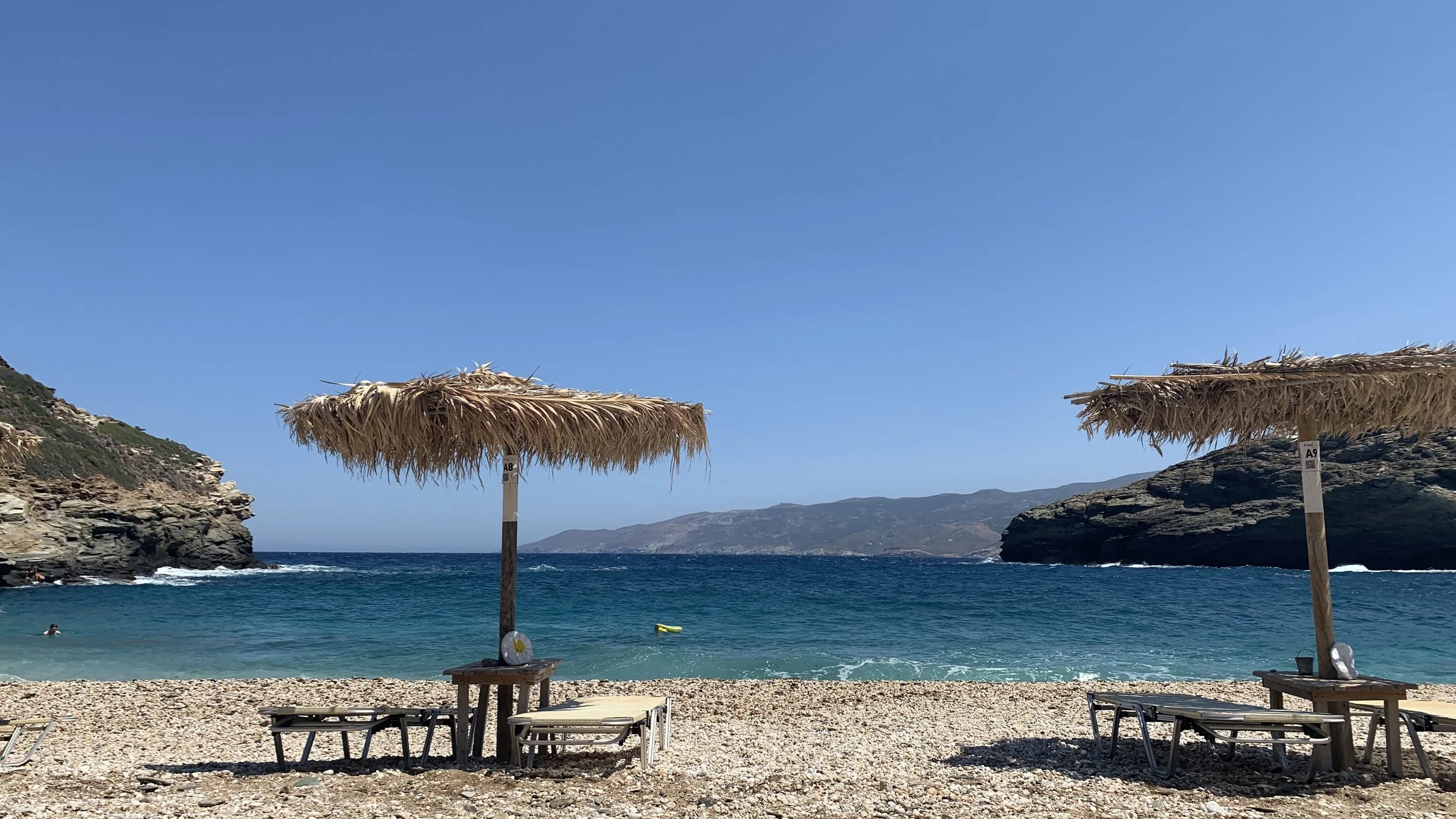 A view of the Andros beach and Greek ocean, with sun loungers and a palm umbrella in front.