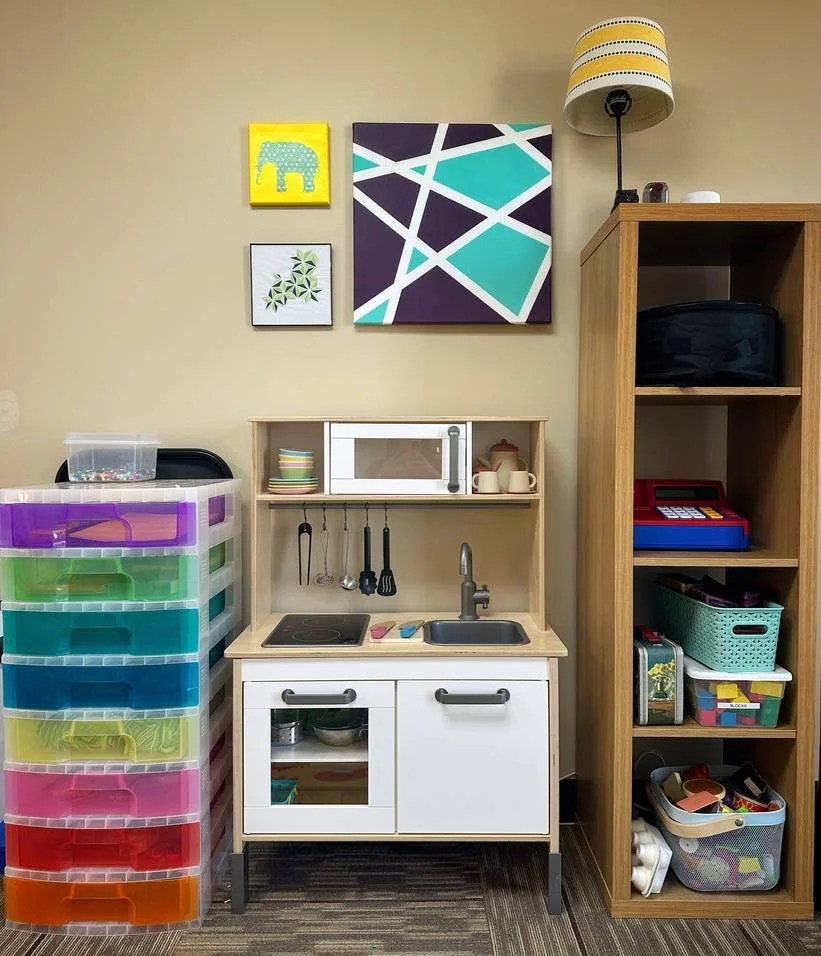 Children's play kitchen with colorful storage drawers on the left, a sink and stovetop in the middle, and a wooden shelf with toy cash register, baskets, and toys on the right. Art hung on the beige wall above.