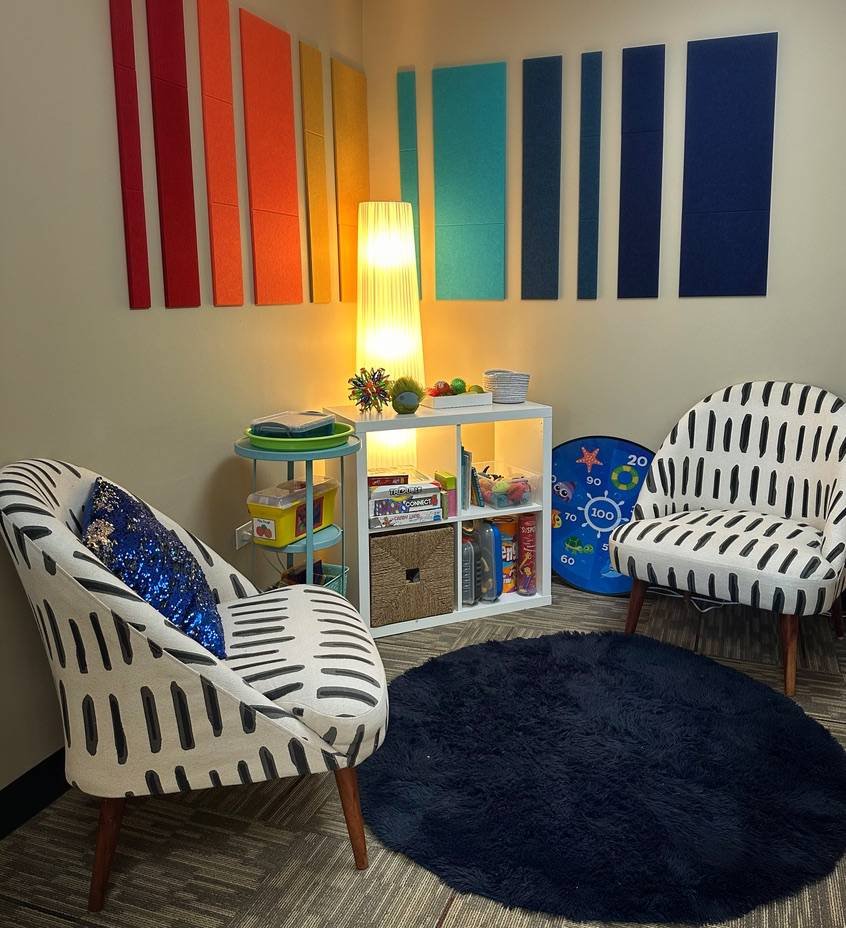 A cozy reading nook with two black and white patterned armchairs, a navy blue rug, a white bookshelf filled with books and toys, a colorful abstract wall art, and a tall floor lamp providing warm lighting.